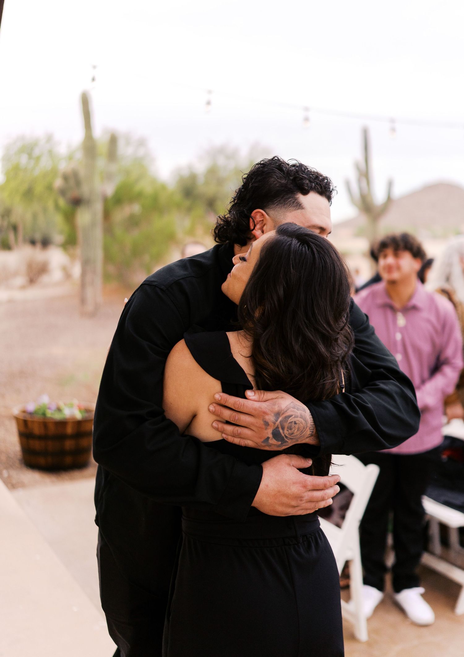 A couple shares a tender embrace while dancing at a desert venue with saguaro cacti in the background.