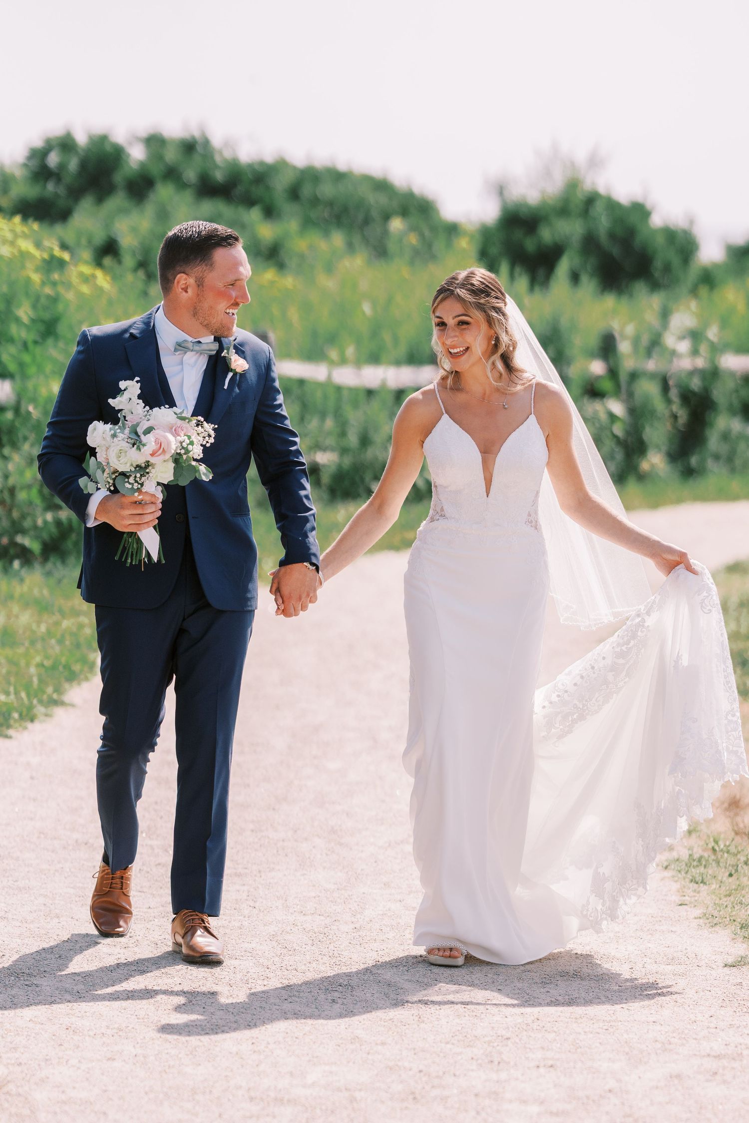 A couple in wedding attire walks together on a sandy beach path during their outdoor summer celebration.