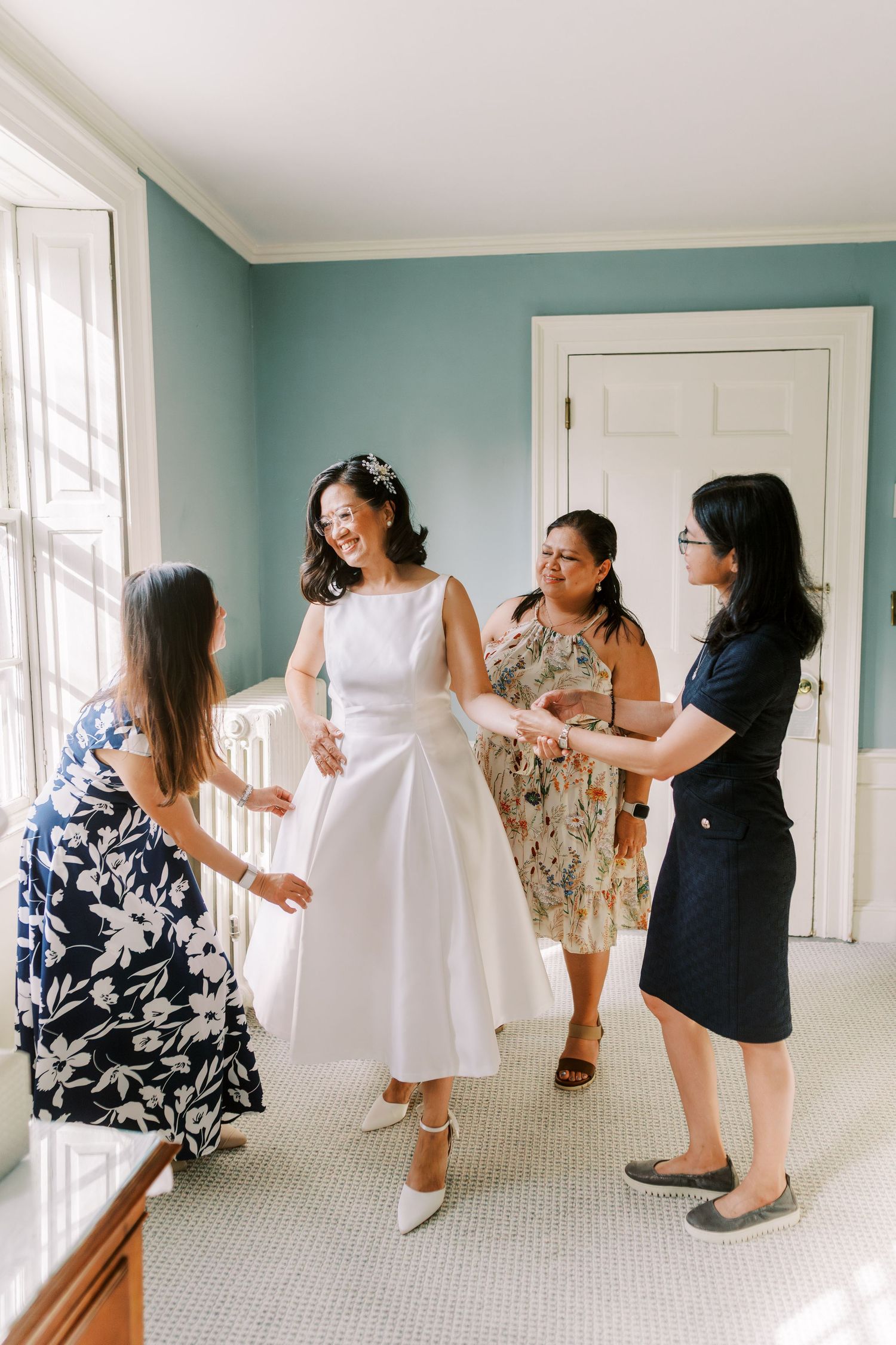 Bridesmaids help with wedding dress preparations in a bright mint-colored room.