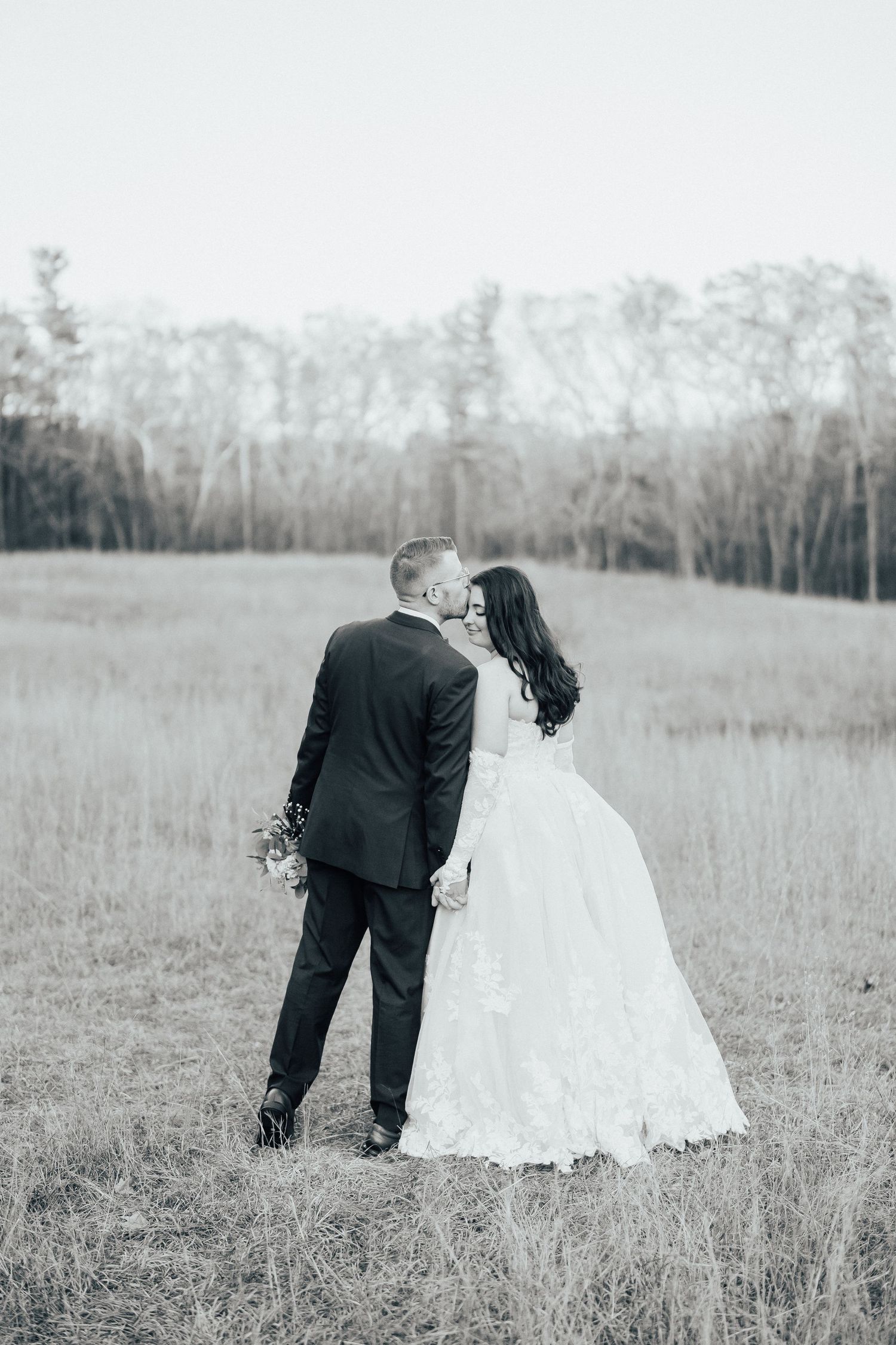 A couple in wedding attire shares a tender moment in a serene field with bare trees in the background.