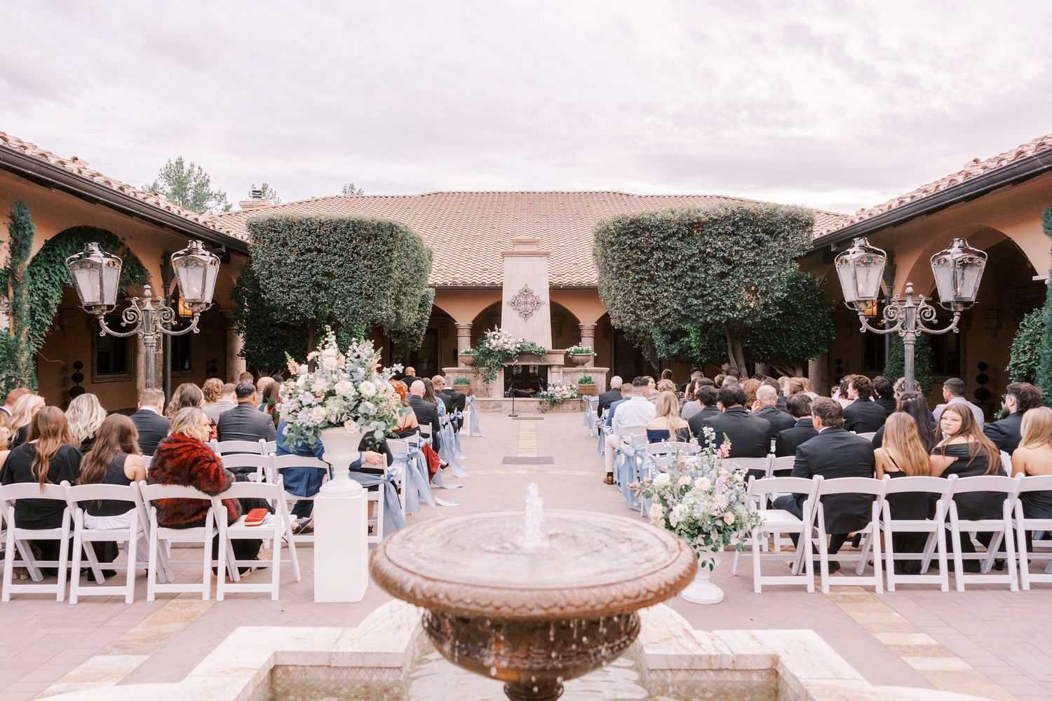 Outdoor wedding venue setup with white chairs, ornate chandeliers, and elegant decor under a covered pavilion.