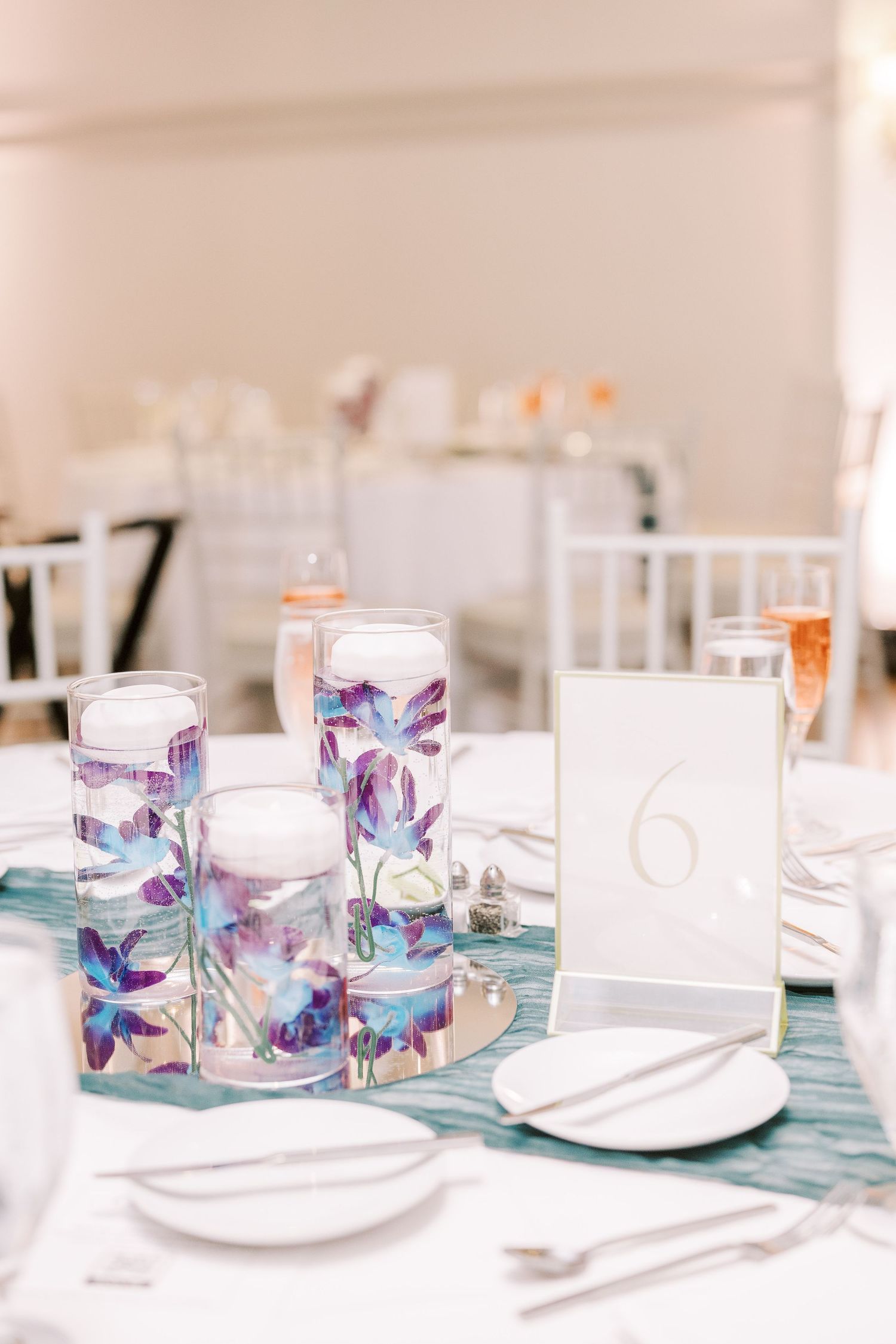 Elegant table setting with blue orchid centerpieces in floating glass cylinders and white place settings for a wedding reception.
