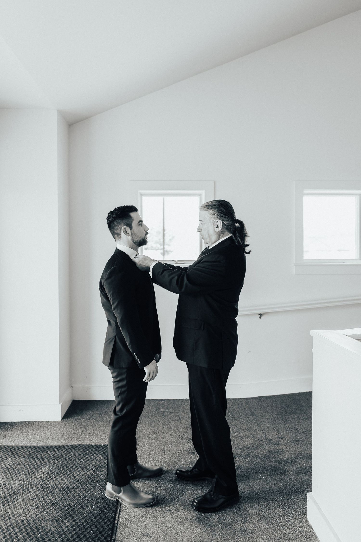 Two people in black suits sharing an intimate moment in a minimalist white room.
