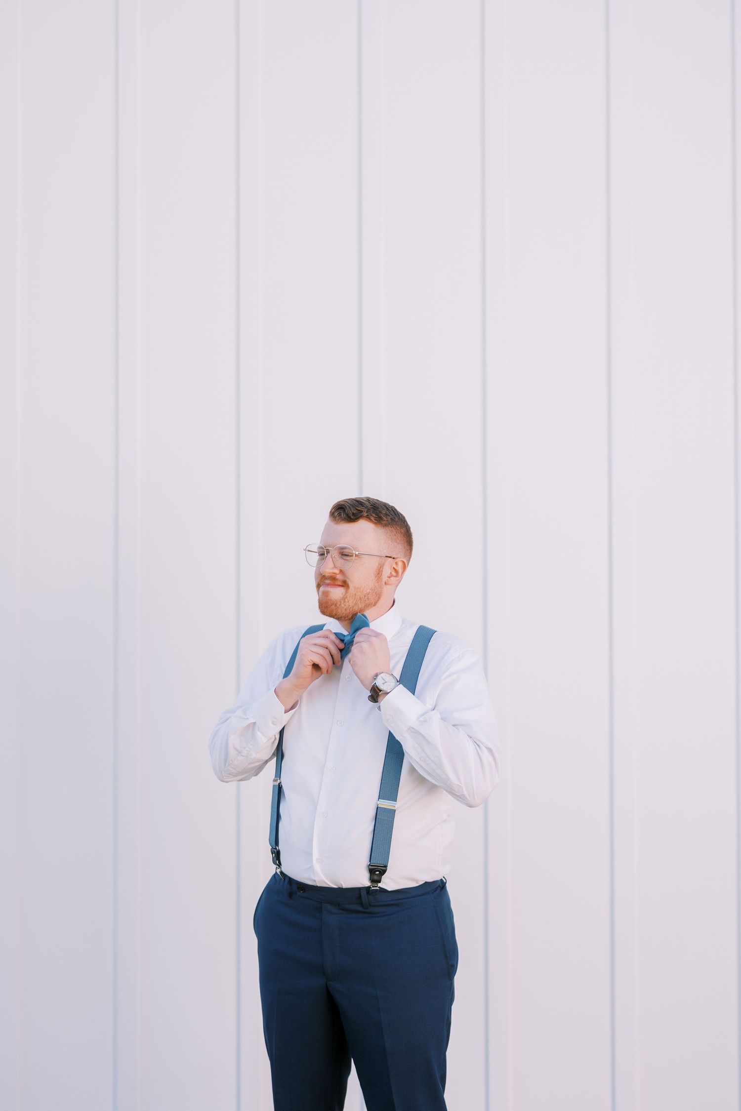 A person wearing a white dress shirt and navy suspenders poses against a white wall in a series of photos.