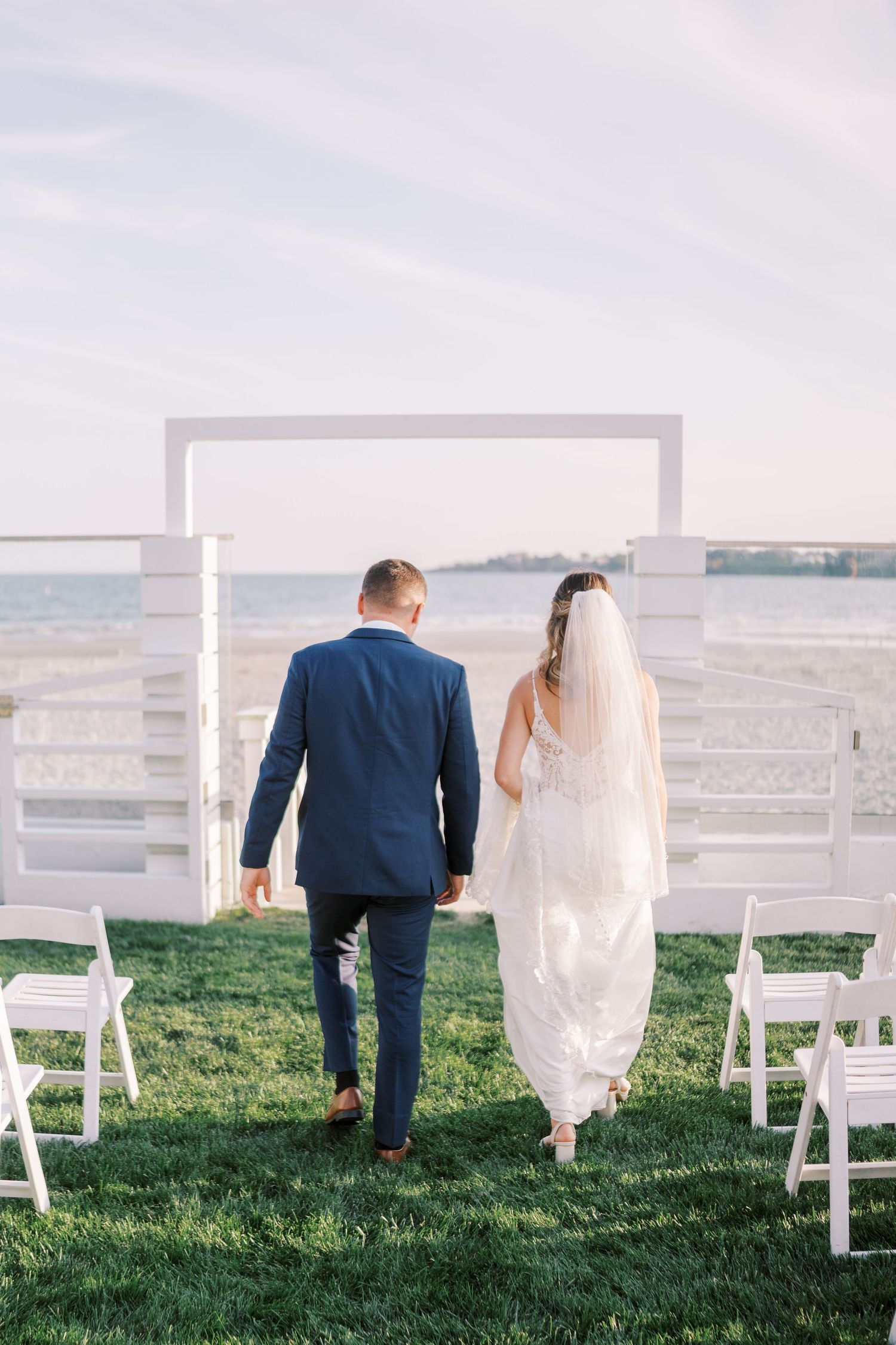 Couple walks through white chairs set up on grassy hill overlooking scenic landscape during outdoor wedding ceremony.