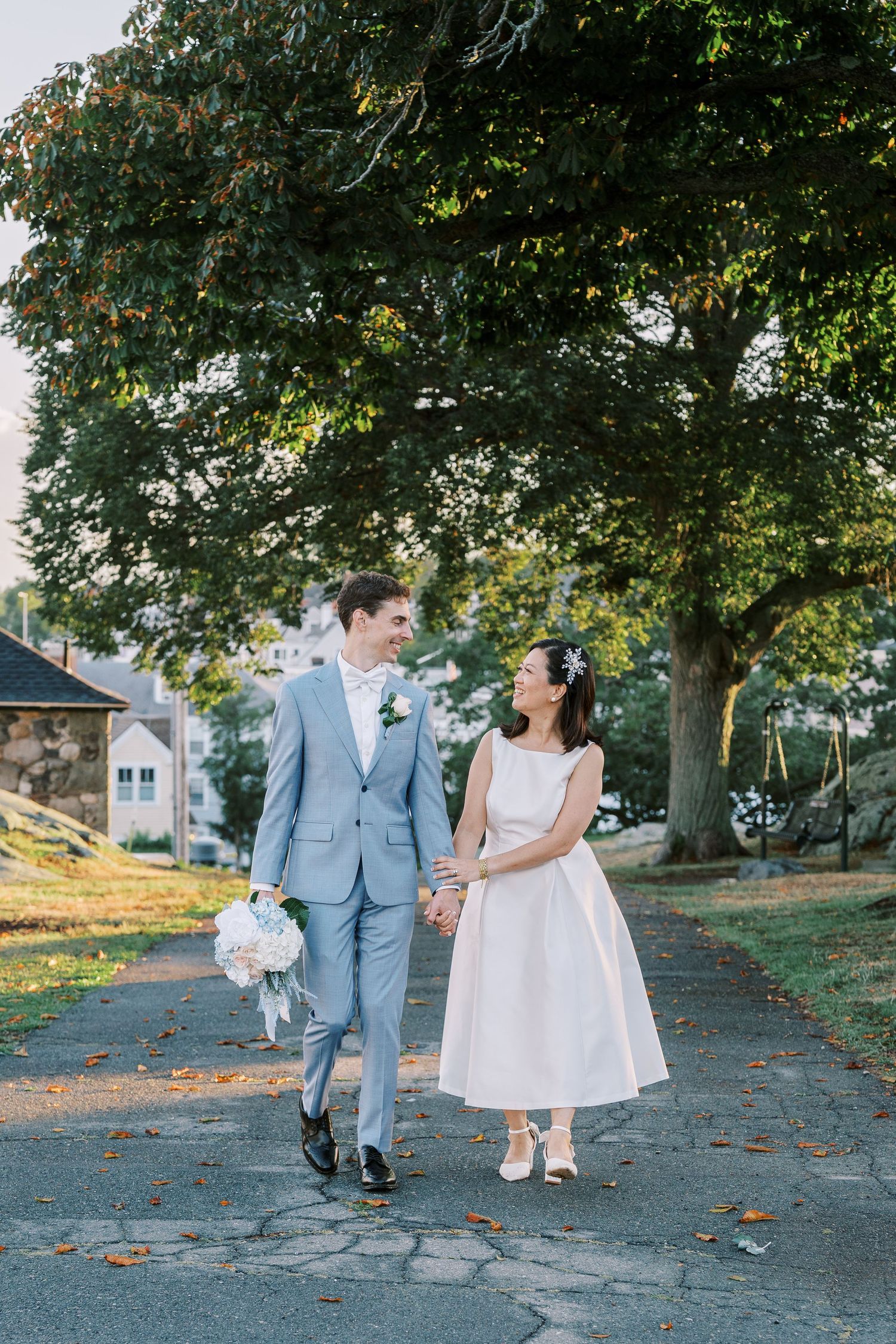 A couple in wedding attire walks along a tree-lined path on their special day, holding hands and smiling at each other.