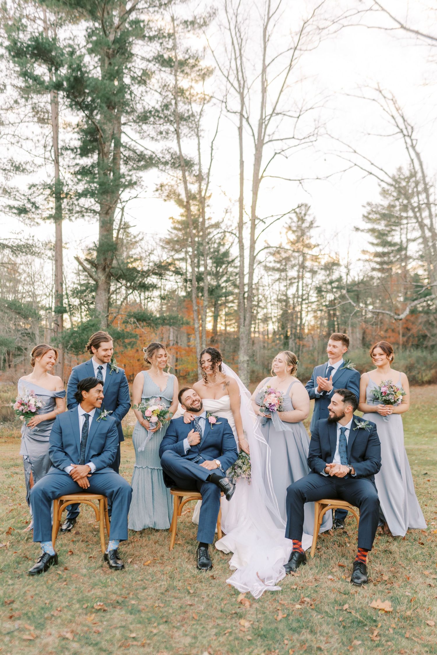 Wedding party in blue suits and gray dresses pose playfully on vintage chairs in an autumn outdoor setting.