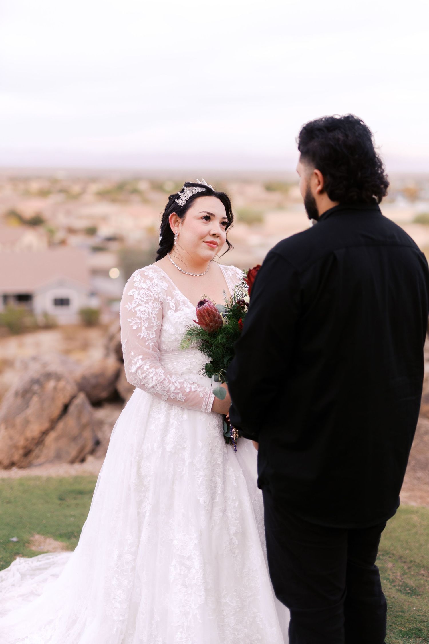 A couple in wedding attire stands outdoors on a hill overlooking desert homes at dusk.