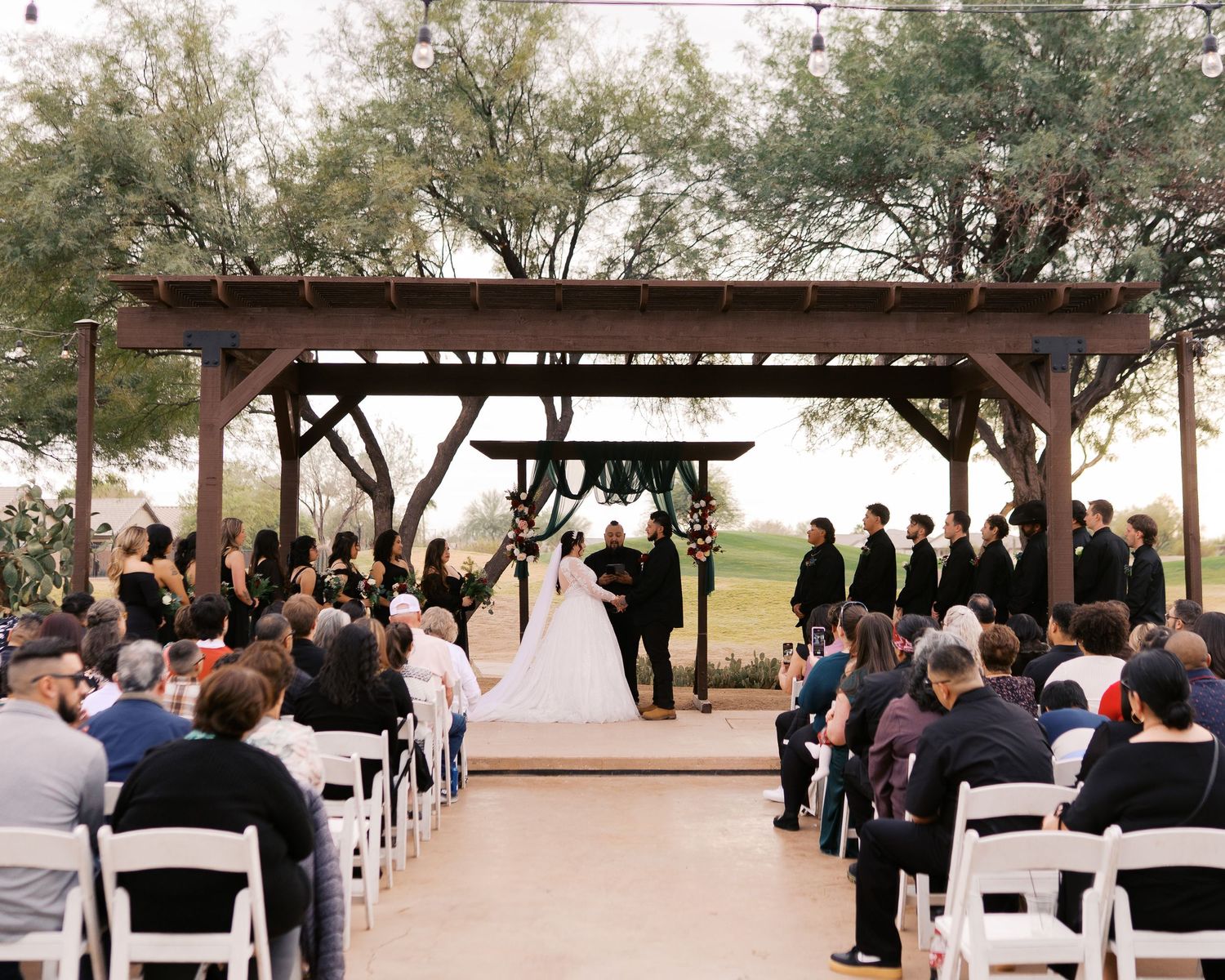 Outdoor wedding ceremony under a wooden pergola with rows of white chairs and guests at a rustic venue during sunset.