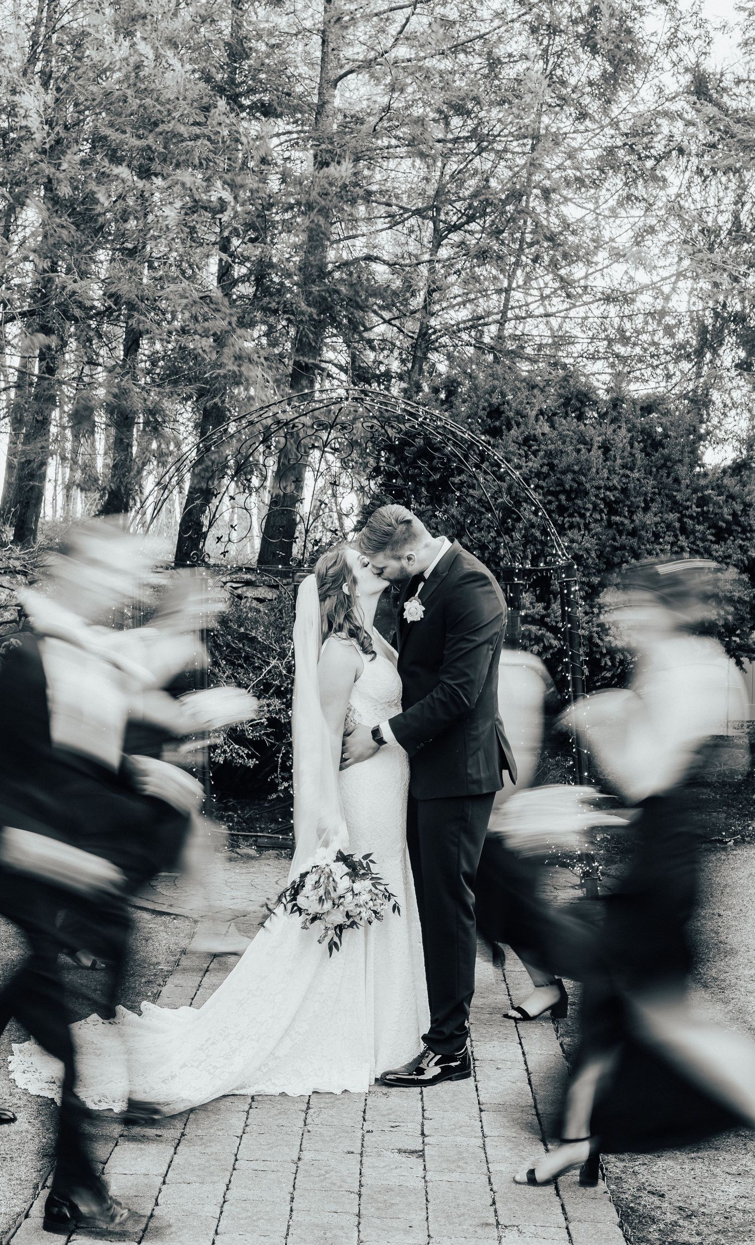 Artistic black and white wedding photo with blurred motion of guests moving around the couple.