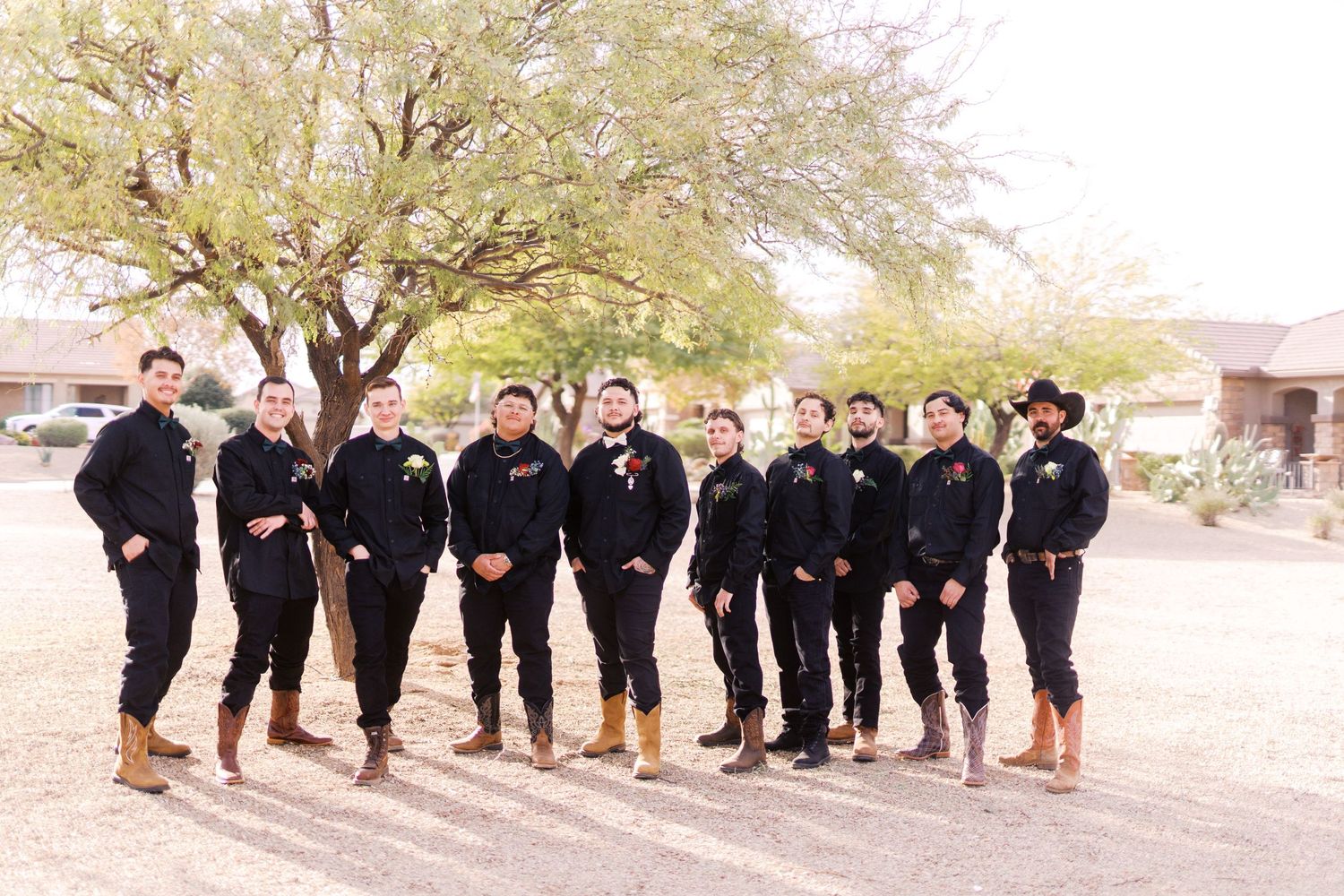 A group wearing matching black outfits and brown boots poses together outdoors under a large tree.