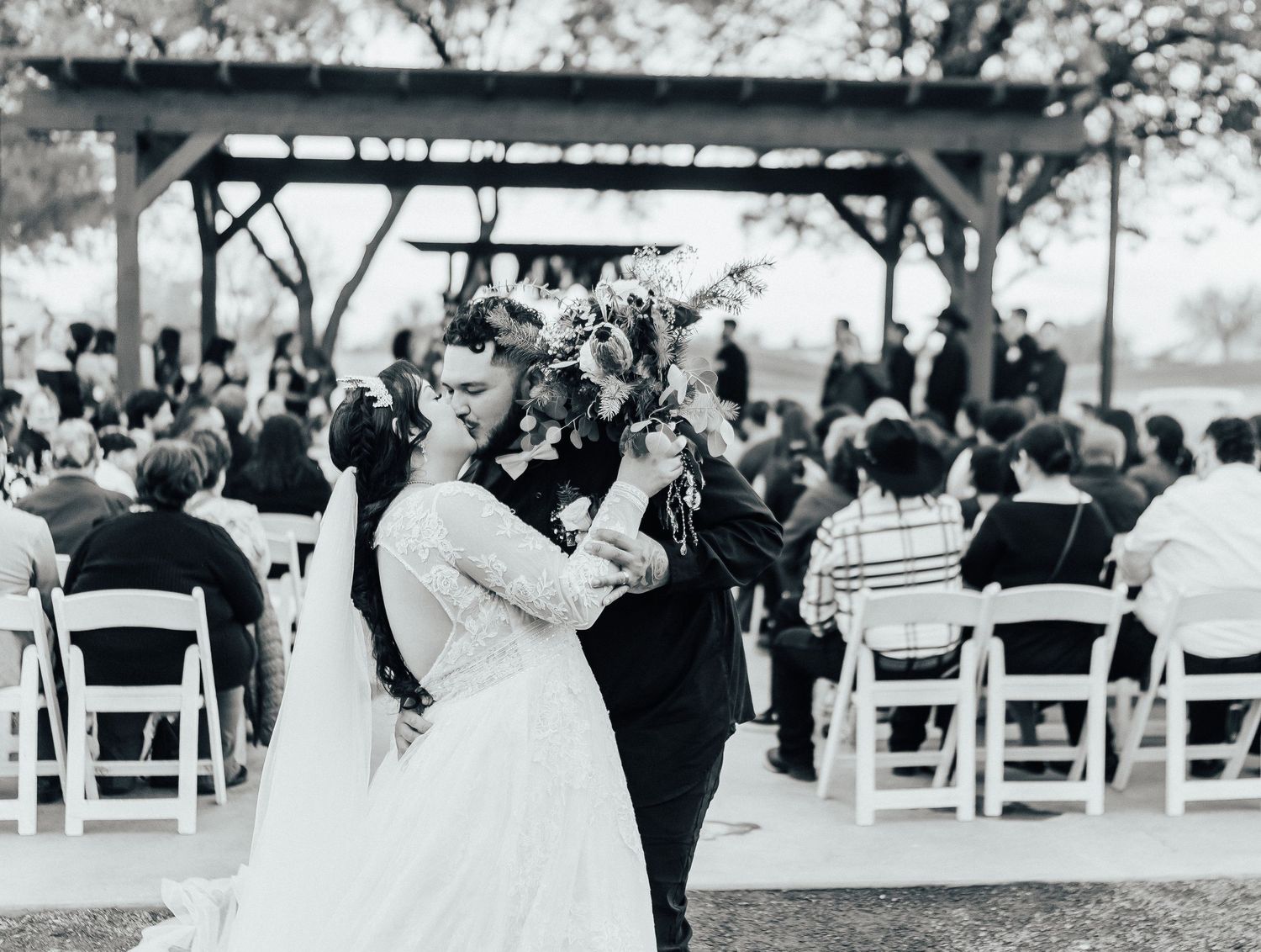 A couple dances at an outdoor wedding ceremony with white chairs and a wooden pergola in the background.