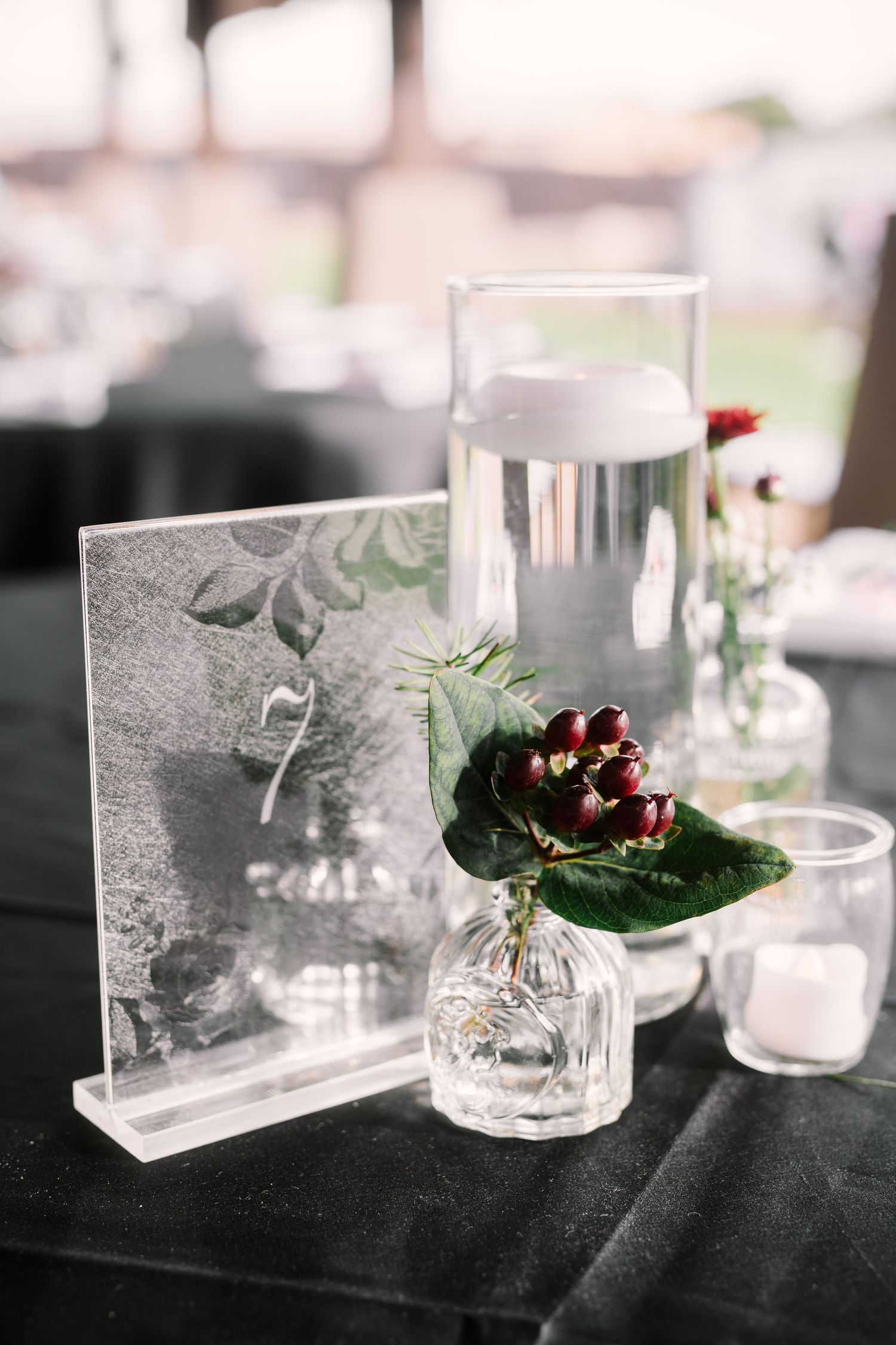 A small glass bud vase with burgundy flower and leaves sits beside a decorative frame on a dark tablecloth.