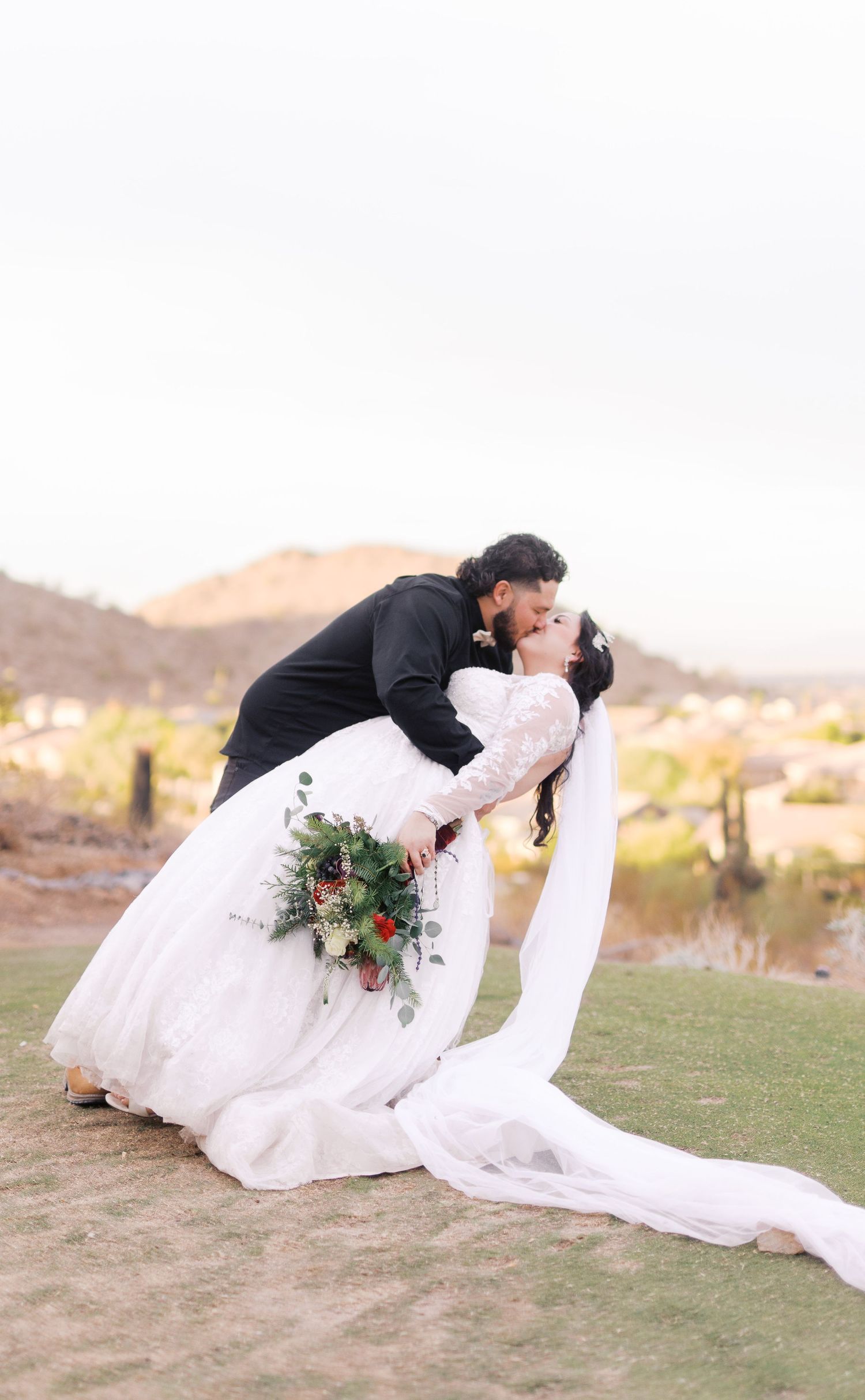 A romantic wedding couple shares a kiss in desert landscape as bride's long white dress train flows across green grass.