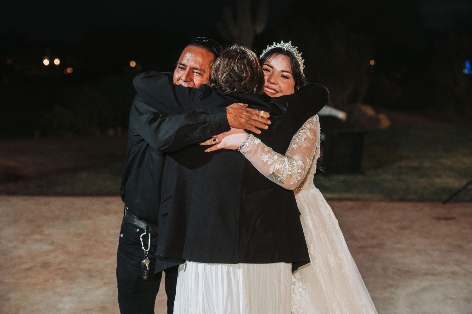 Three people share a joyful embrace at an outdoor nighttime celebration with dark lighting and formal attire.