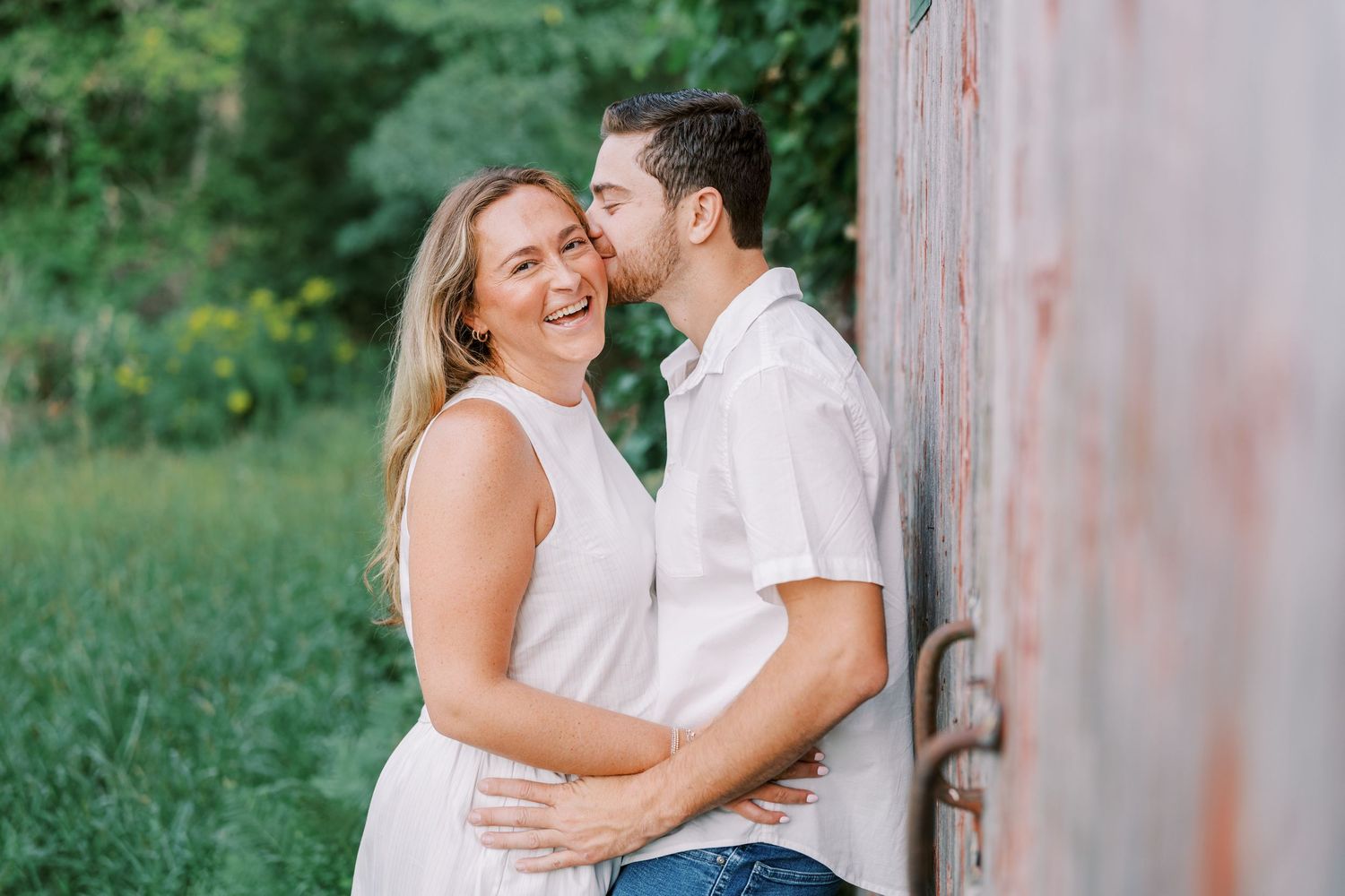 A couple in coordinating white outfits sharing an intimate moment against a rustic wall outdoors.