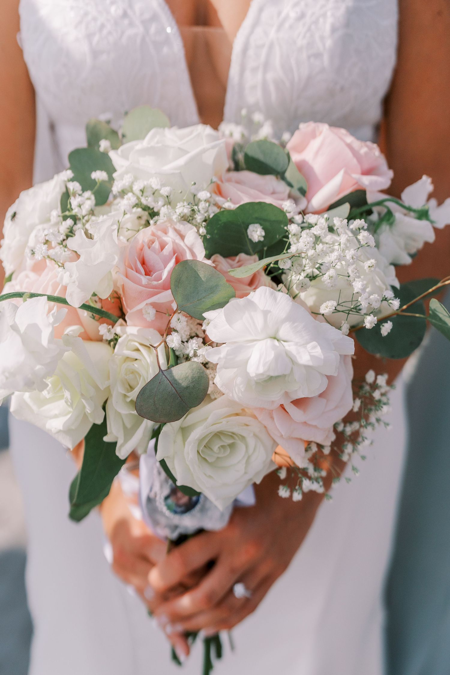Elegant bridal bouquet with white and blush pink roses, baby's breath and eucalyptus leaves held against white wedding dress.