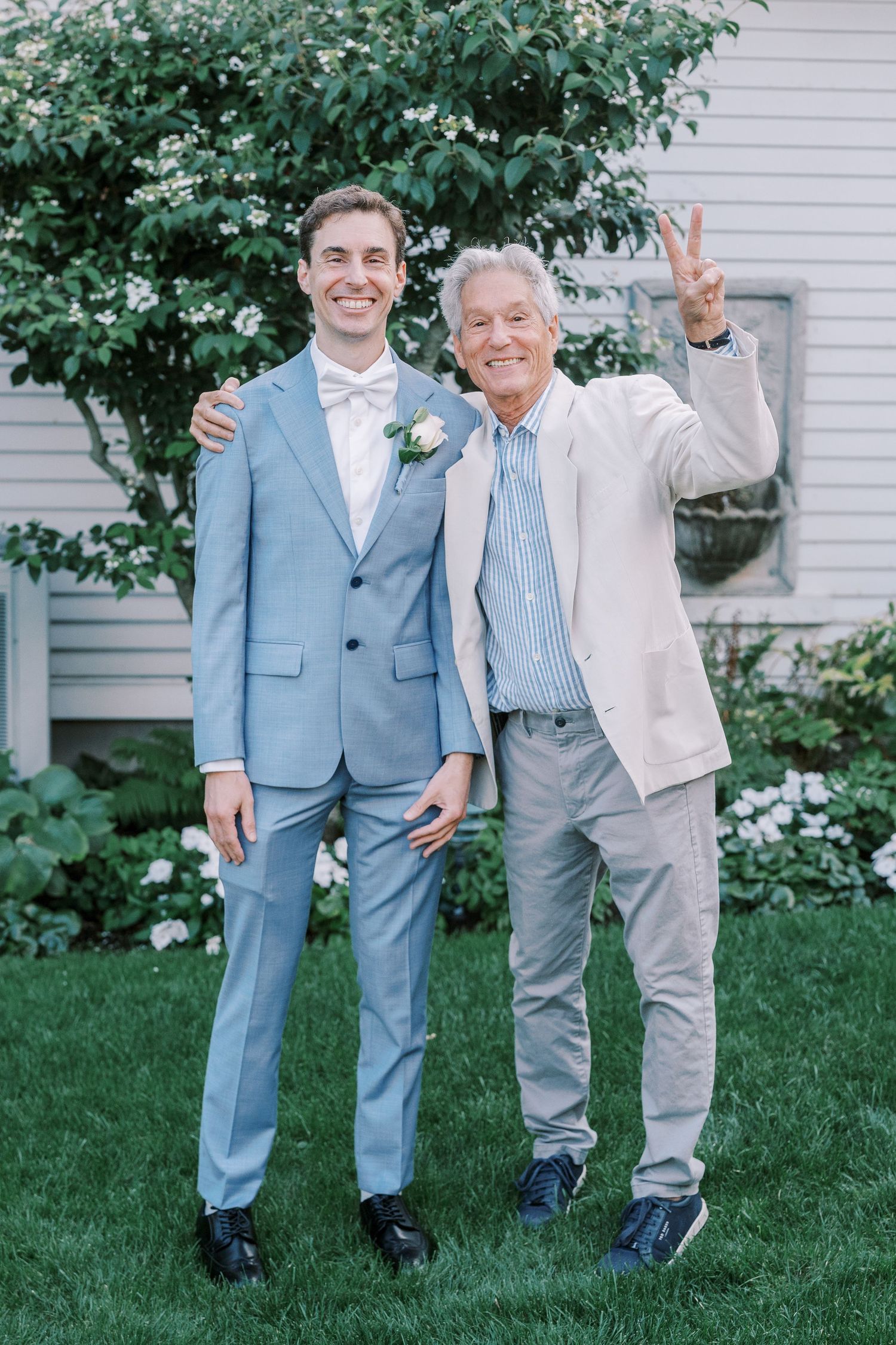 Two people in light and blue suits posing together on grass with a white house in background.