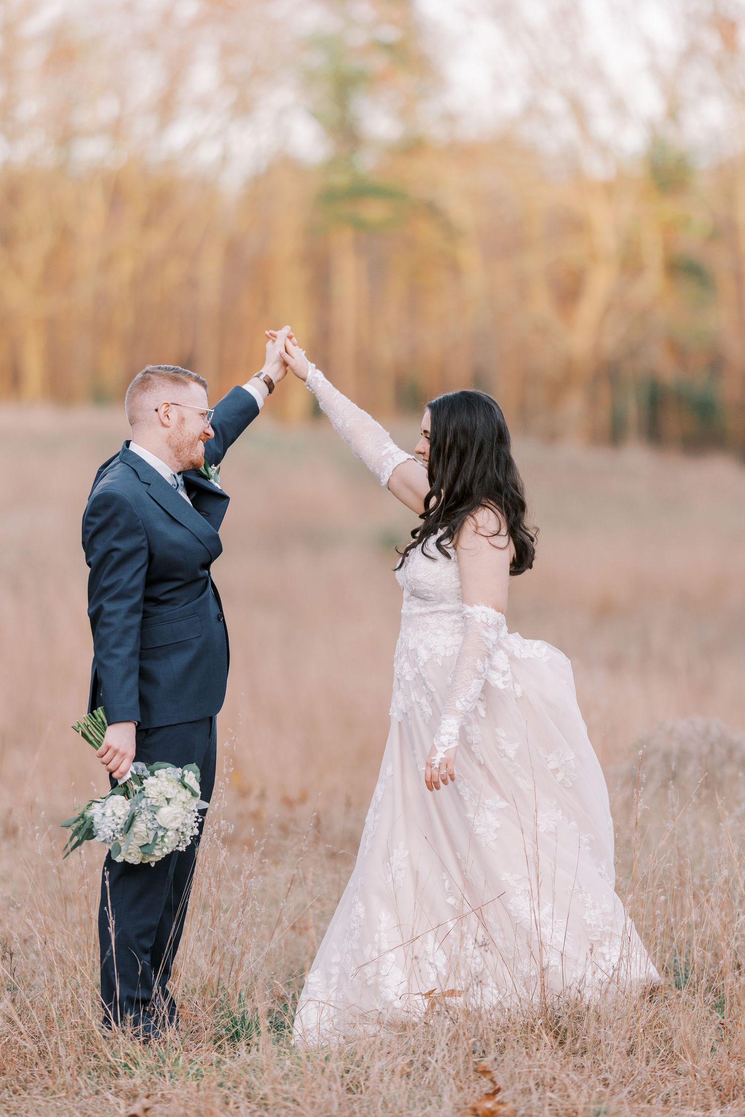 A couple in wedding attire dances and twirls joyfully together in a golden autumn field at sunset.