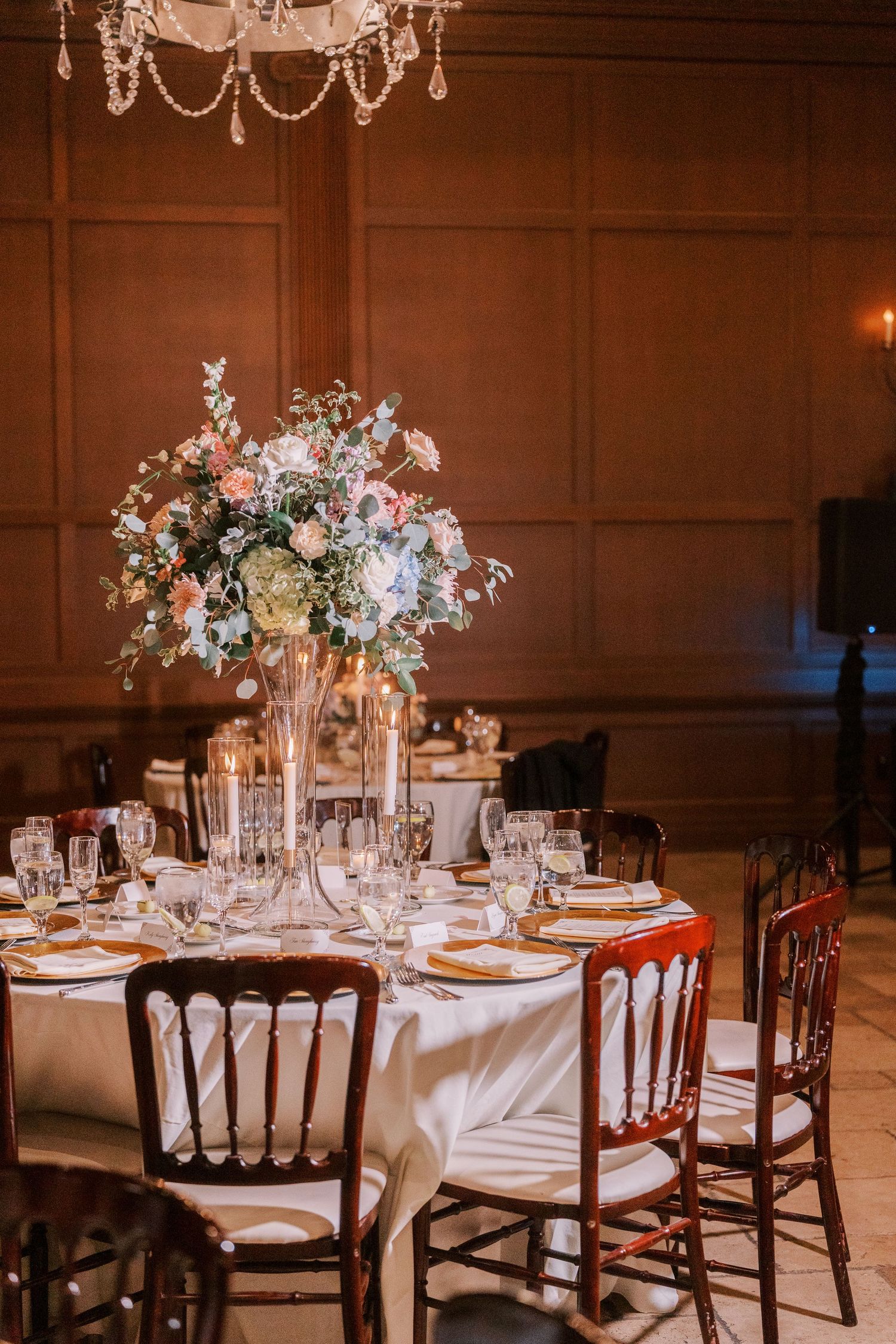 Elegant wedding reception tables with white linens, floral centerpieces, and mahogany chiavari chairs in a warm-lit ballroom.