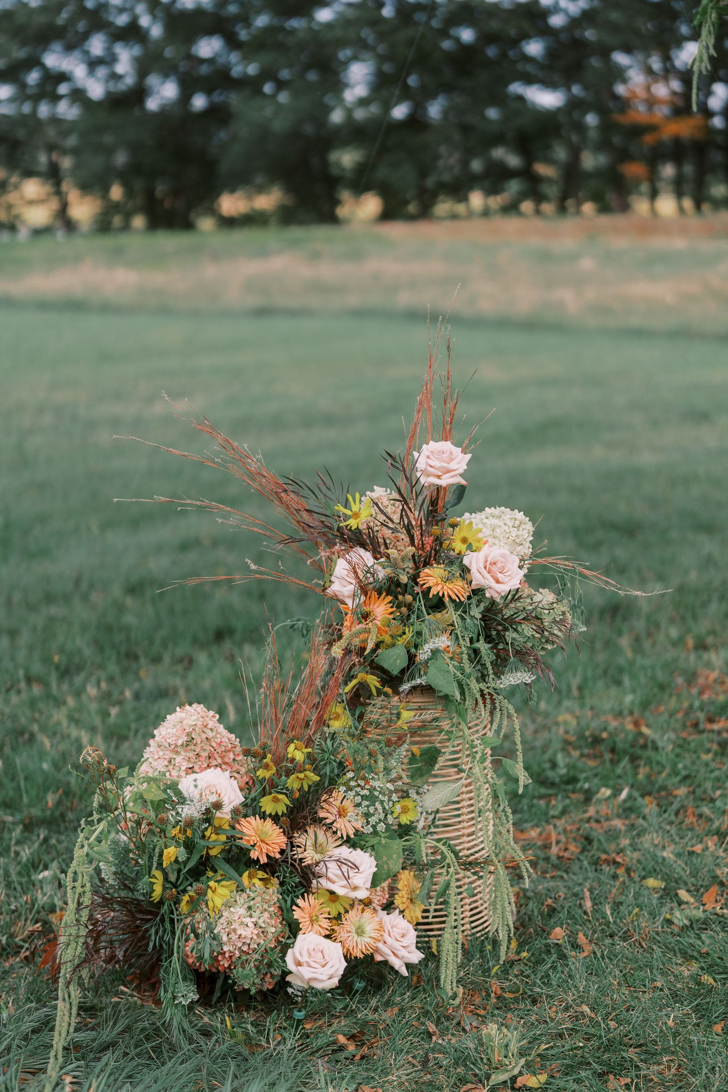 Rustic floral arrangements with pink roses and dried elements displayed on grass at an outdoor wedding.