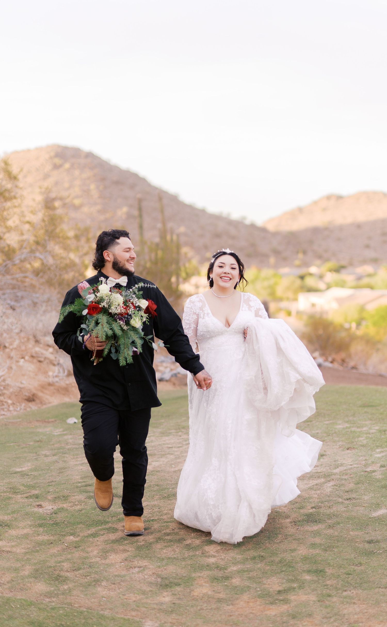 A joyful wedding couple runs hand in hand across desert terrain with mountains in the background at sunset.