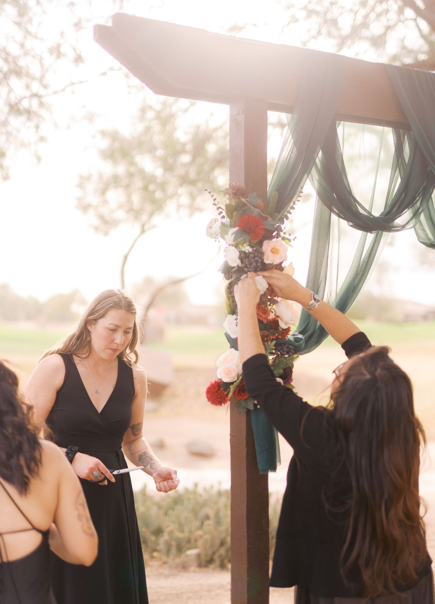 People decorate an outdoor wooden arbor with floral arrangements and green fabric draping in a sunlit setting.