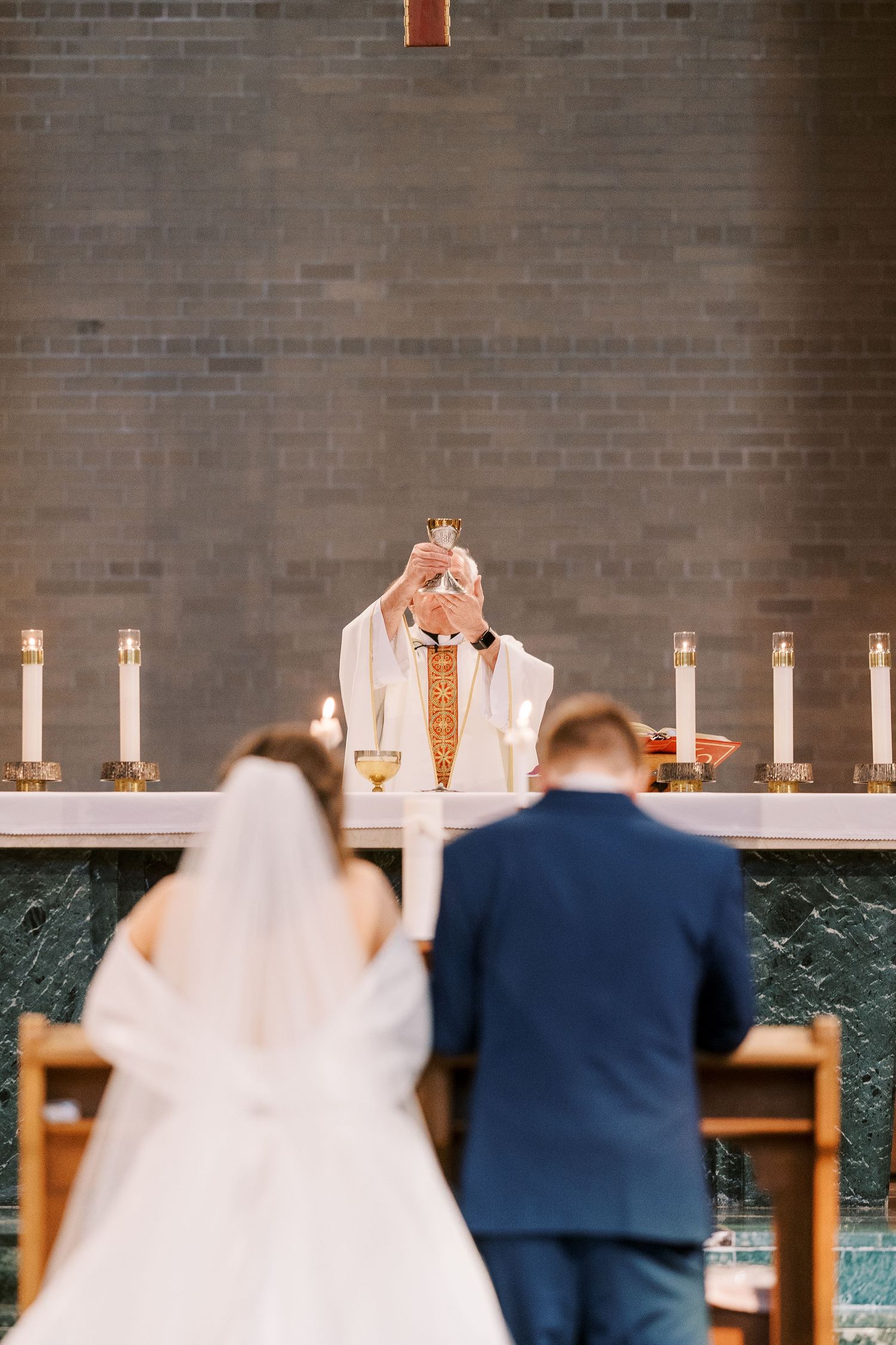 A couple in wedding attire stands before lit candles in a church with exposed brick walls.