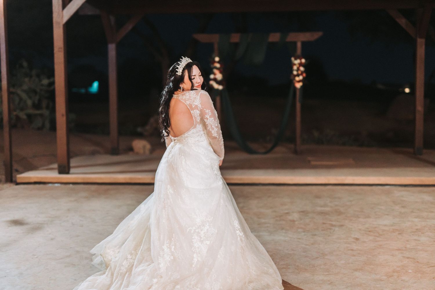 A bride in a flowing lace wedding dress stands with her back turned in a rustic venue with hanging lights.