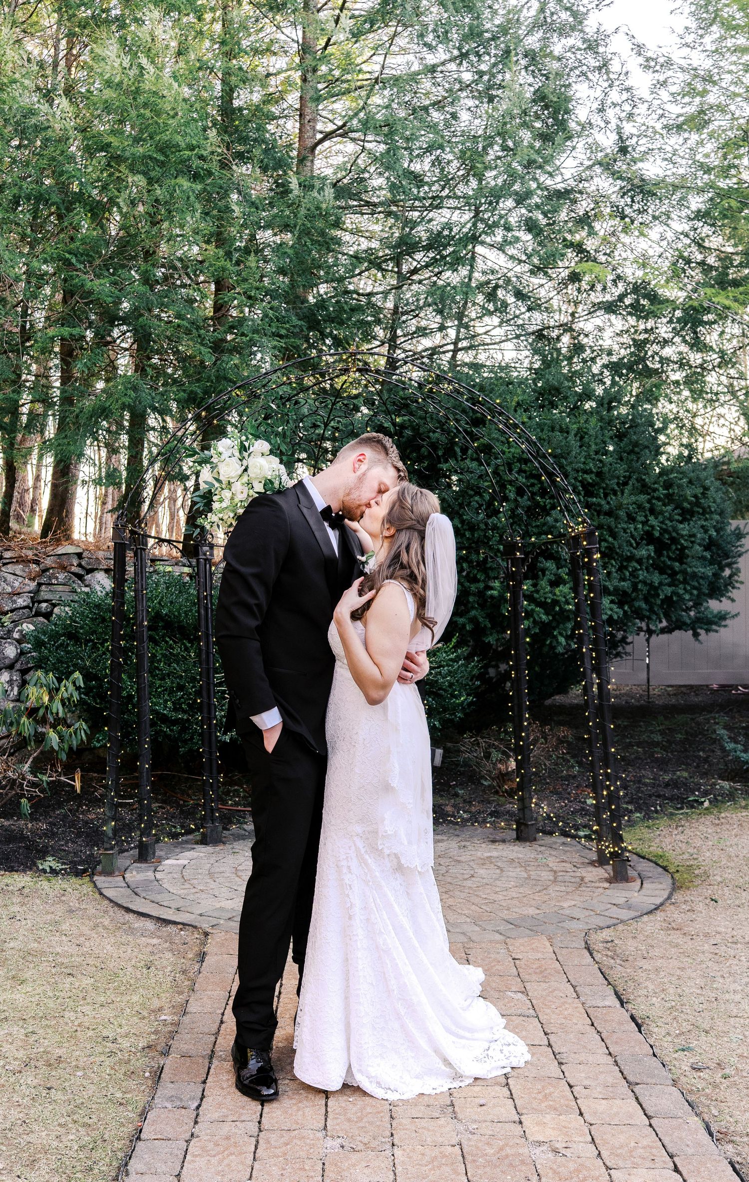 A romantic moment between newlyweds sharing a kiss on a brick path surrounded by lush greenery.