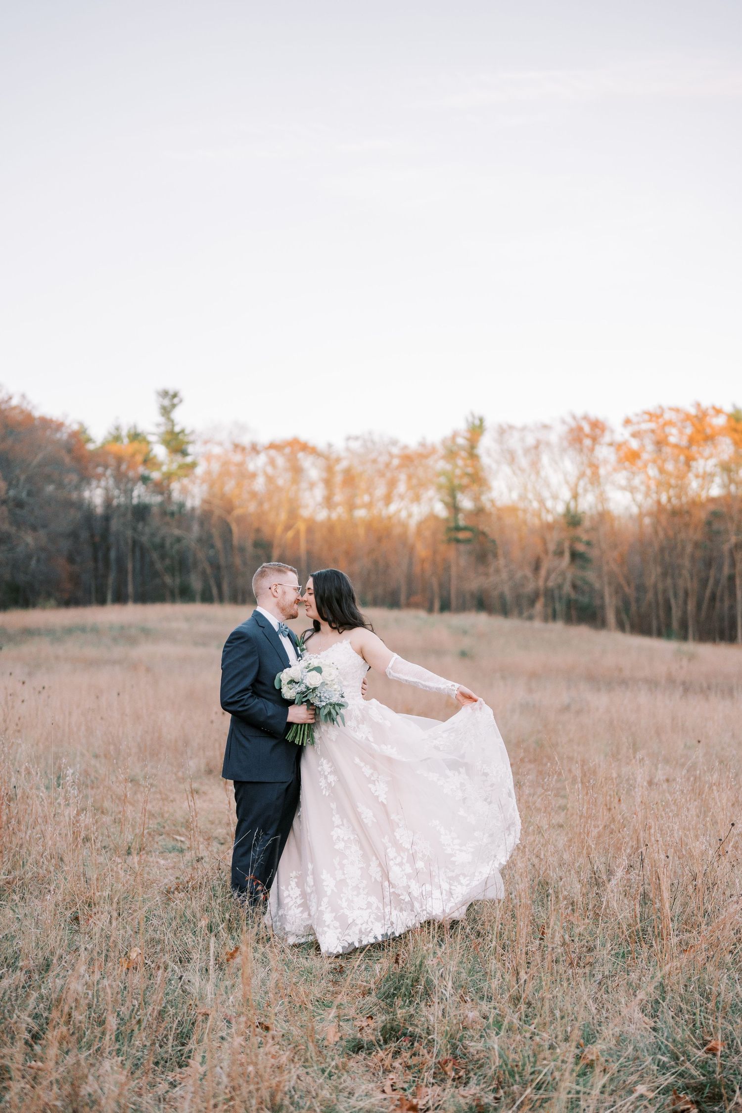 Romantic wedding couple embraces in a golden autumn field at sunset with trees in the background.