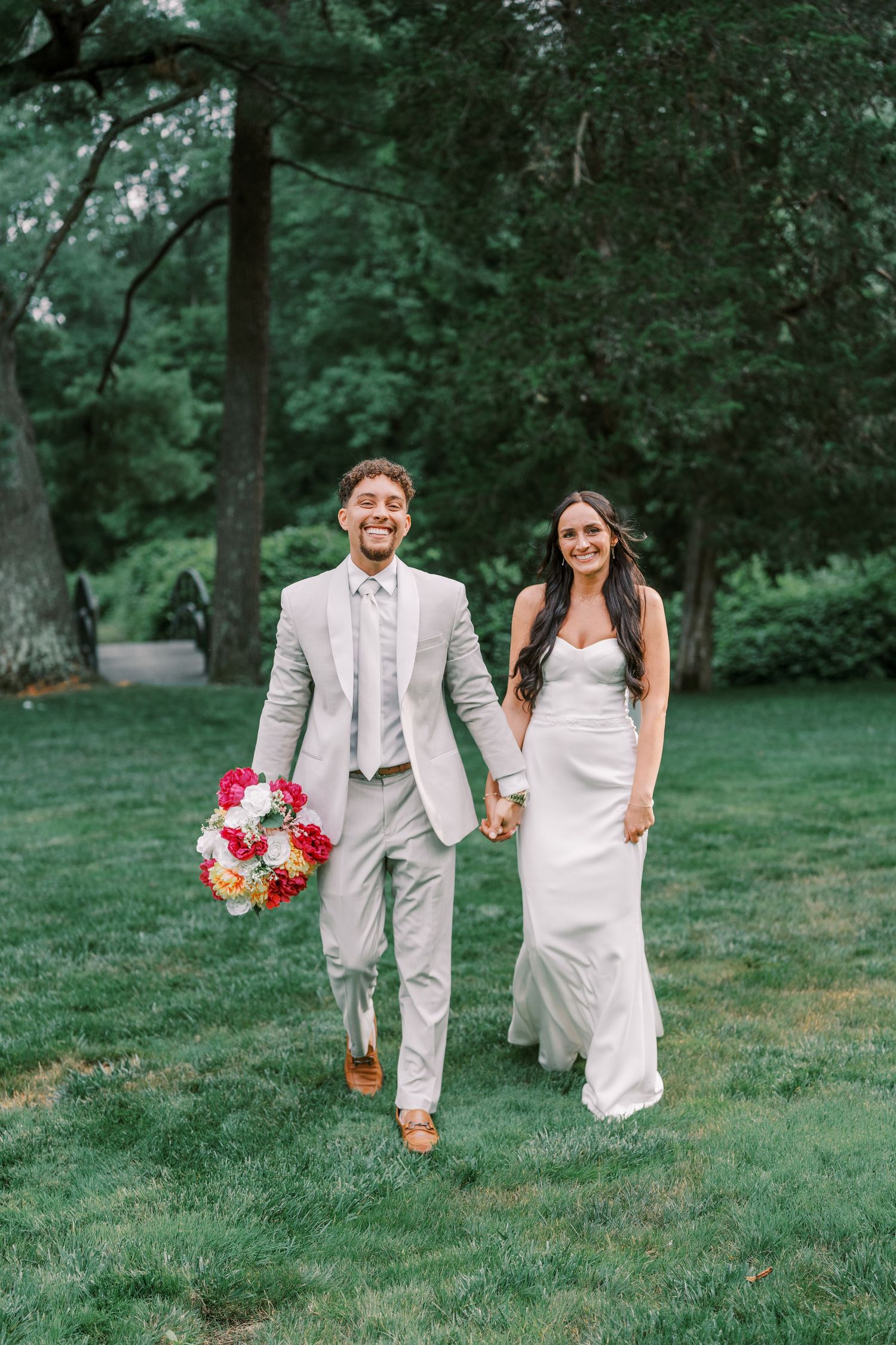 A wedding couple holding colorful bouquets walks together on lush green grass in a park setting.