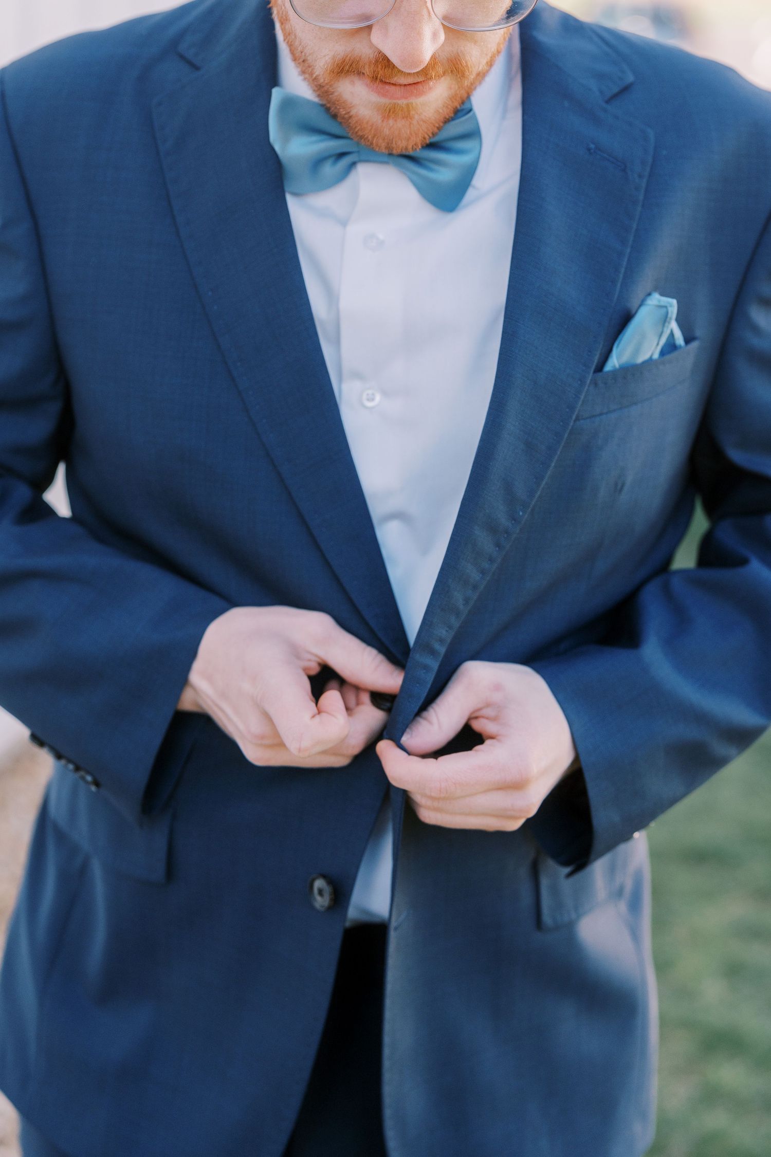 A navy suit jacket with light blue bow tie and matching pocket square being buttoned by the wearer.
