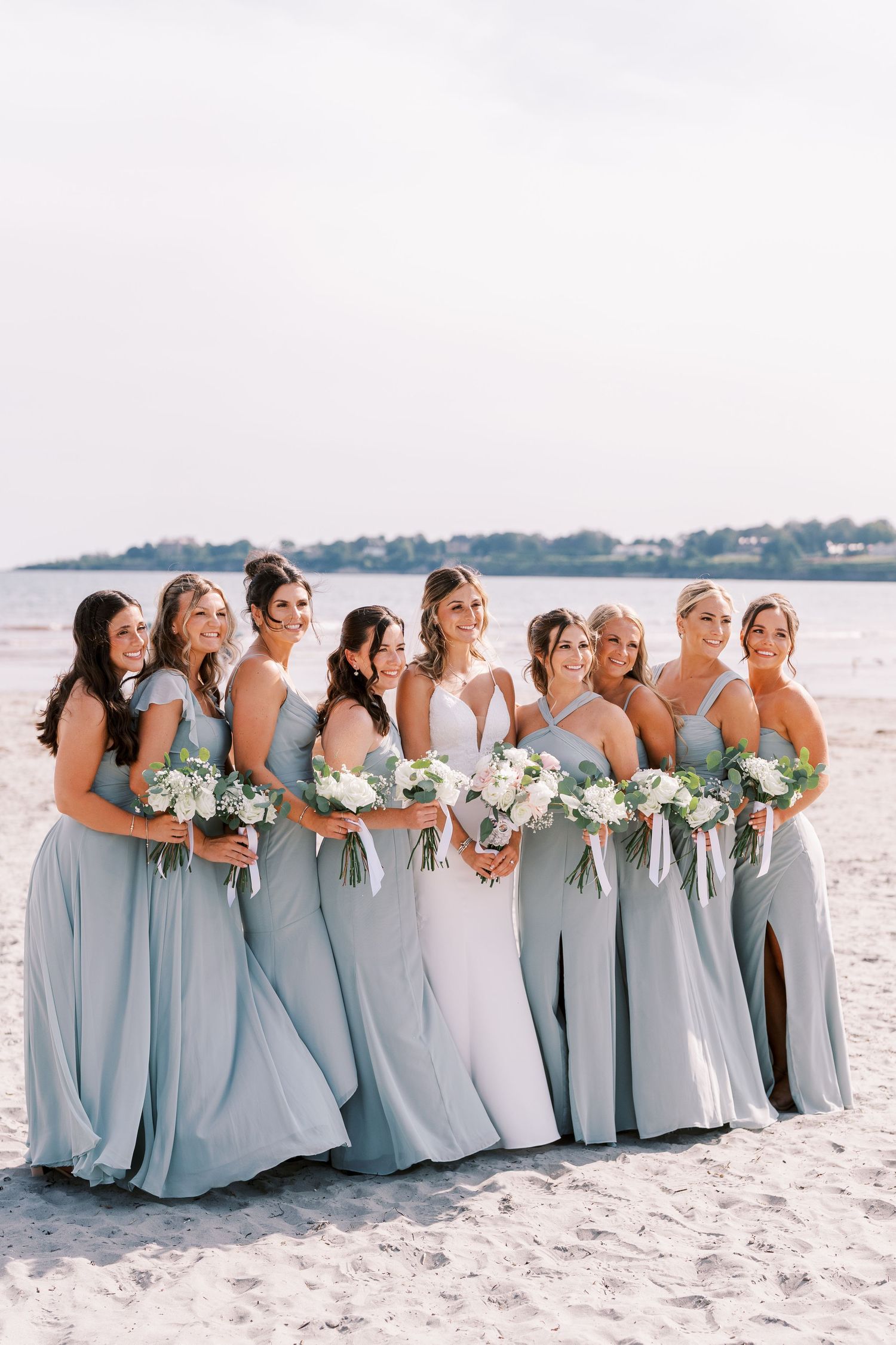A row of bridesmaids in light blue dresses stand together on a sunny beach holding white floral bouquets.