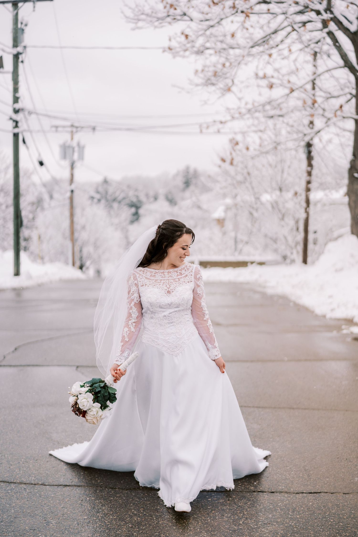 A bride in a long-sleeved lace wedding gown poses on a snowy winter day with bare trees in the background.