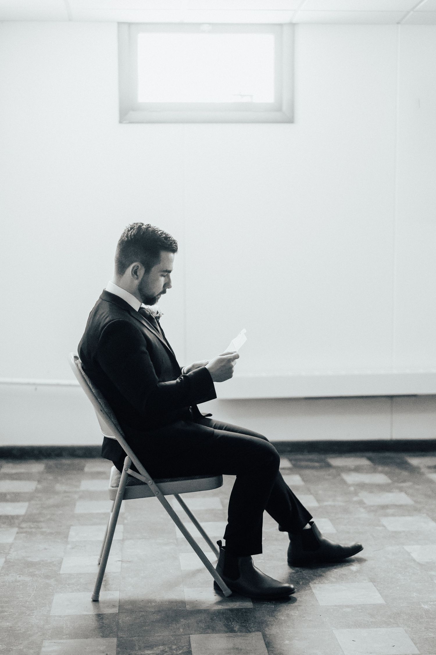 Man in black suit sitting alone in minimalist white room reading during quiet moment.