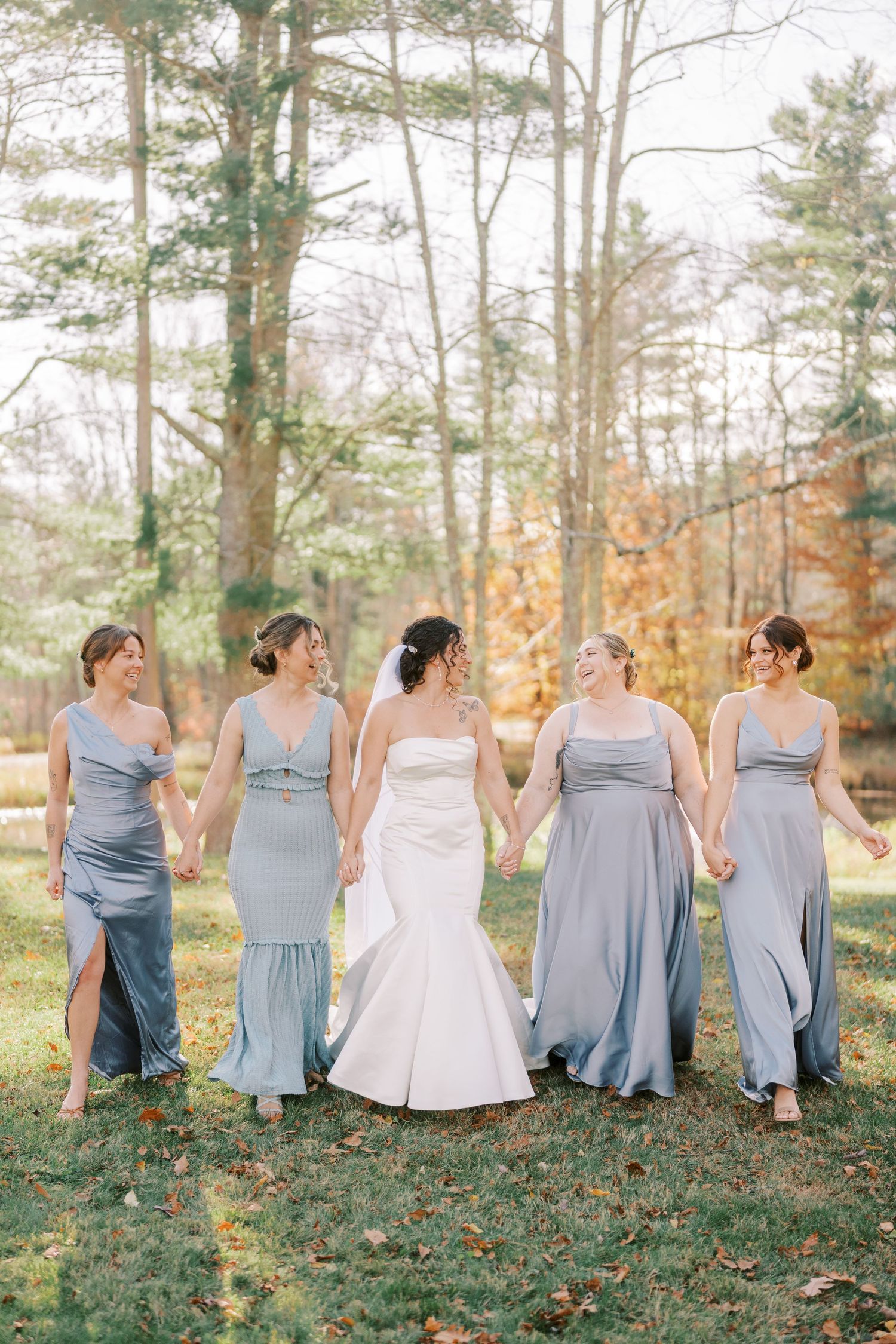 A wedding party in dusty blue bridesmaid dresses poses together on grass during an autumn outdoor photoshoot.