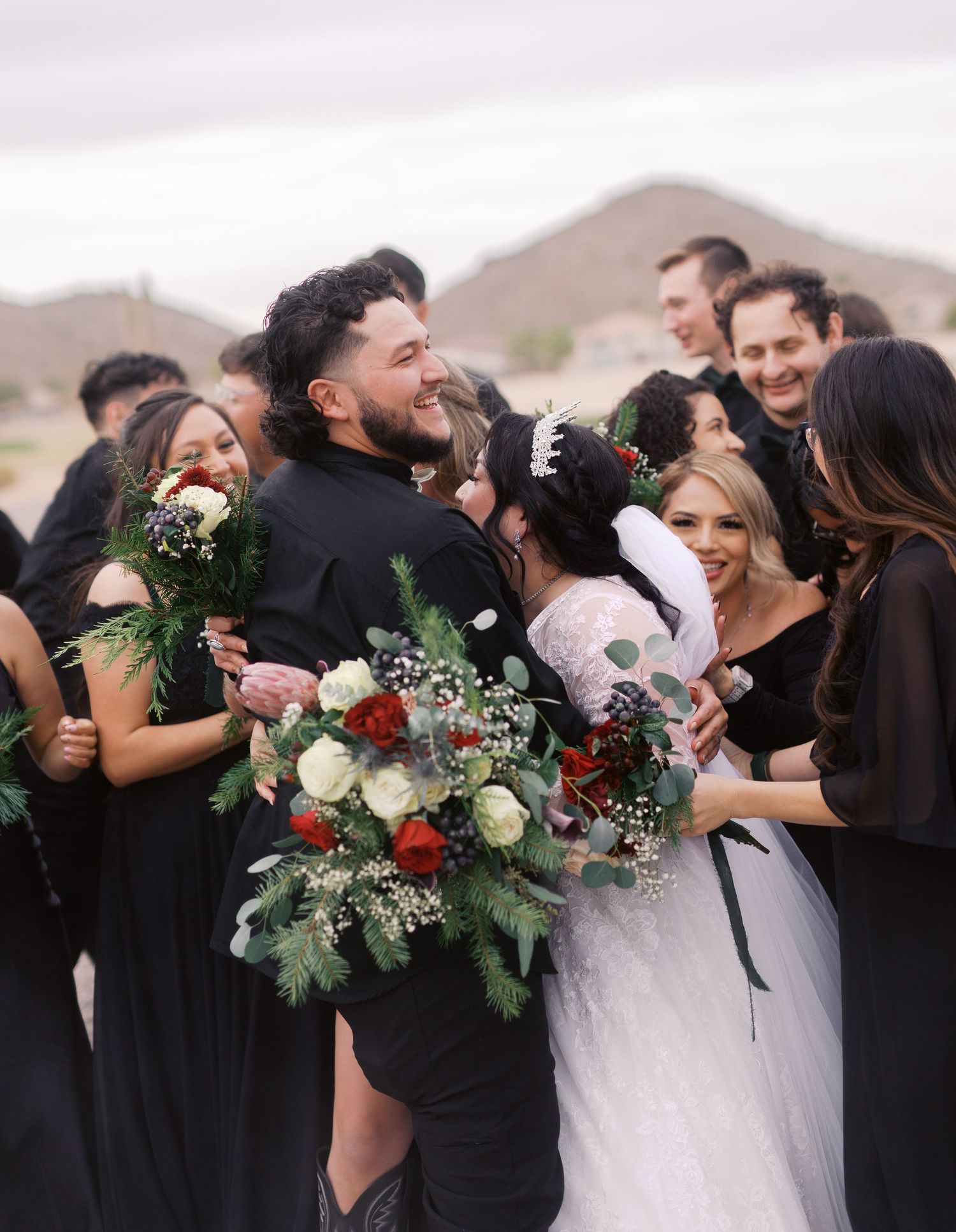 Wedding party laughs together in black attire with bride holding bouquet of red and white flowers against mountain backdrop.