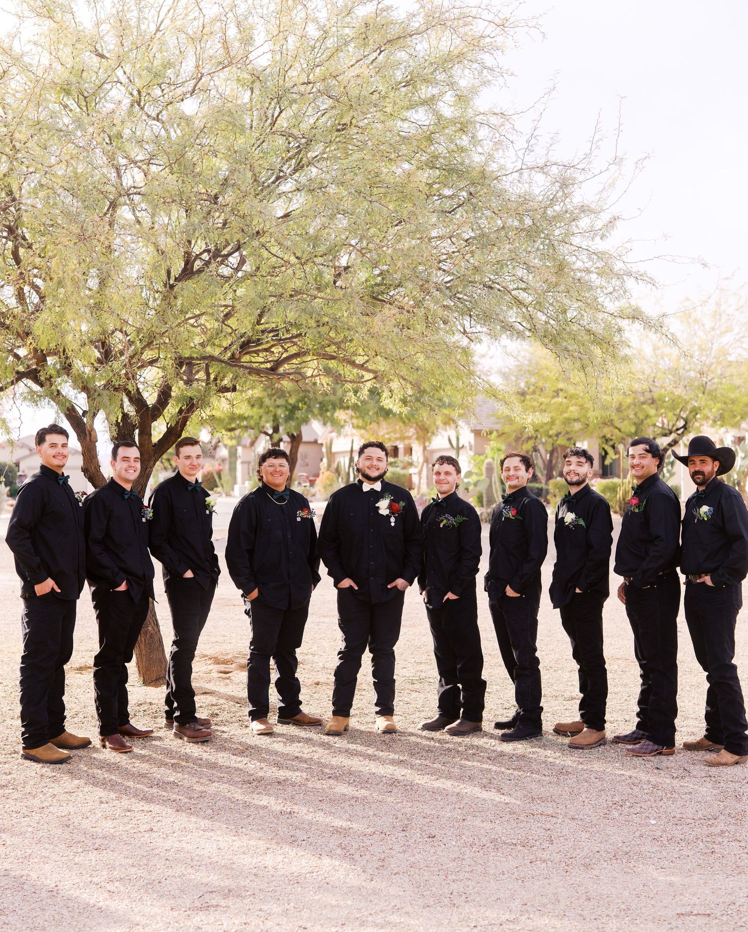 A line of groomsmen dressed in black suits pose together under a large desert tree on a dirt path.