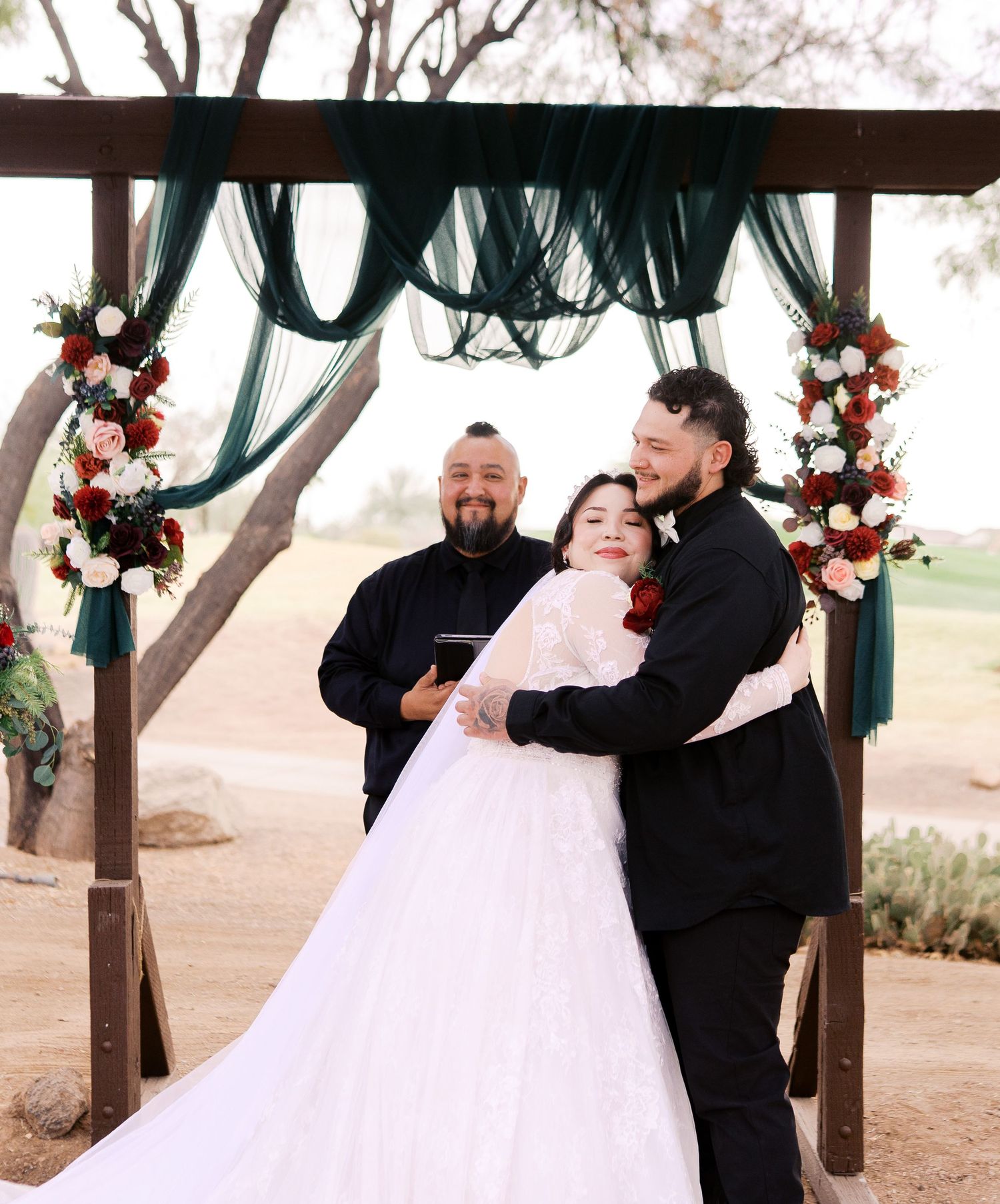 A wedding ceremony taking place under a wooden arch decorated with emerald green draping and coral floral arrangements.