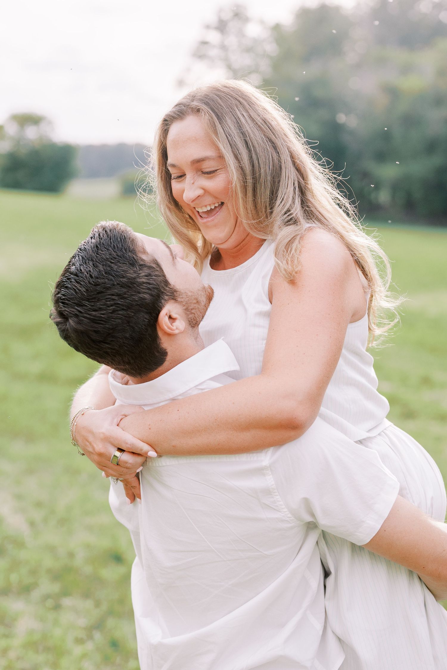 Playful couple in white clothing embracing and laughing in a sunny field.