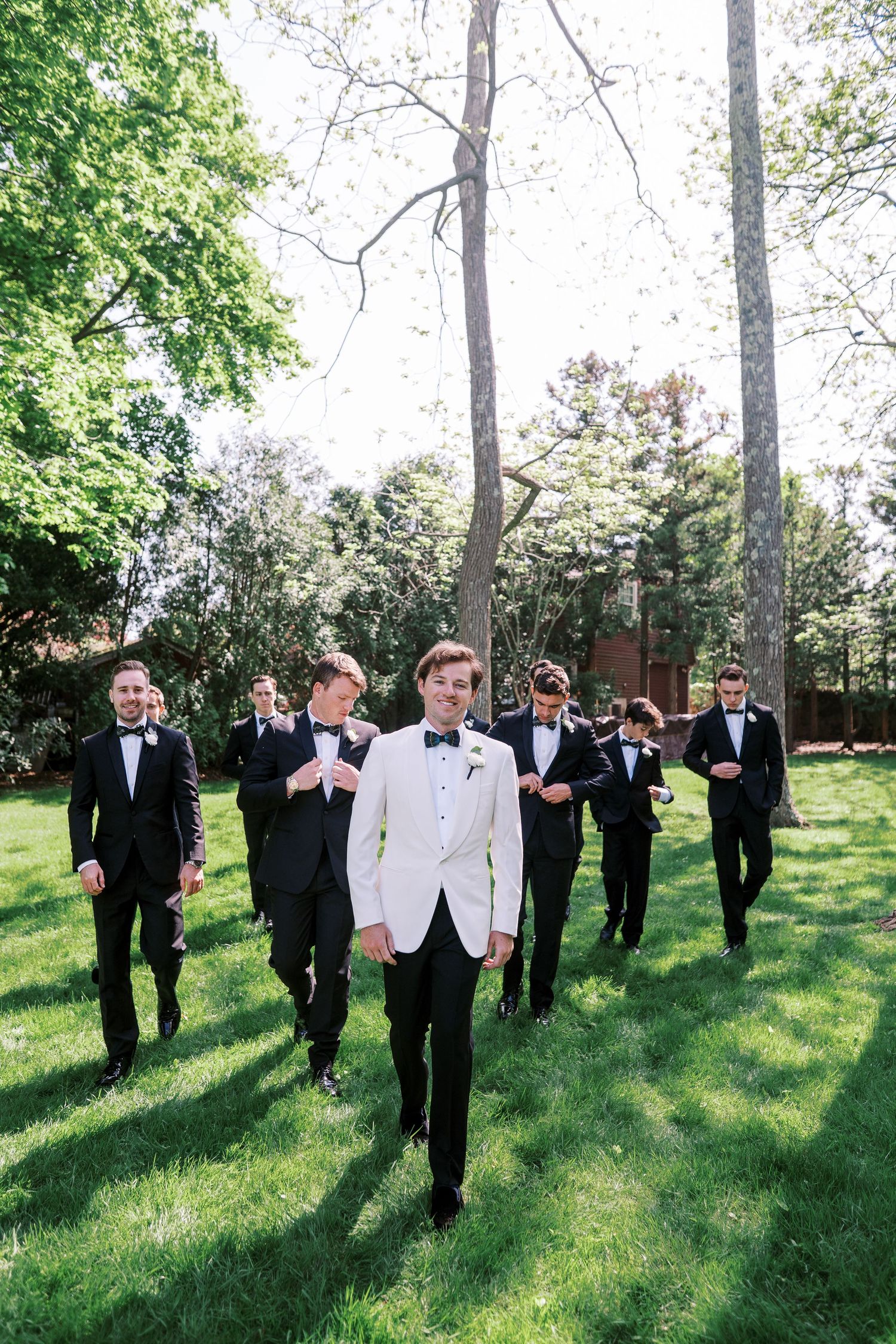 Groomsmen in black tuxedos walking outdoors on grass with one person wearing a white dinner jacket in front.
