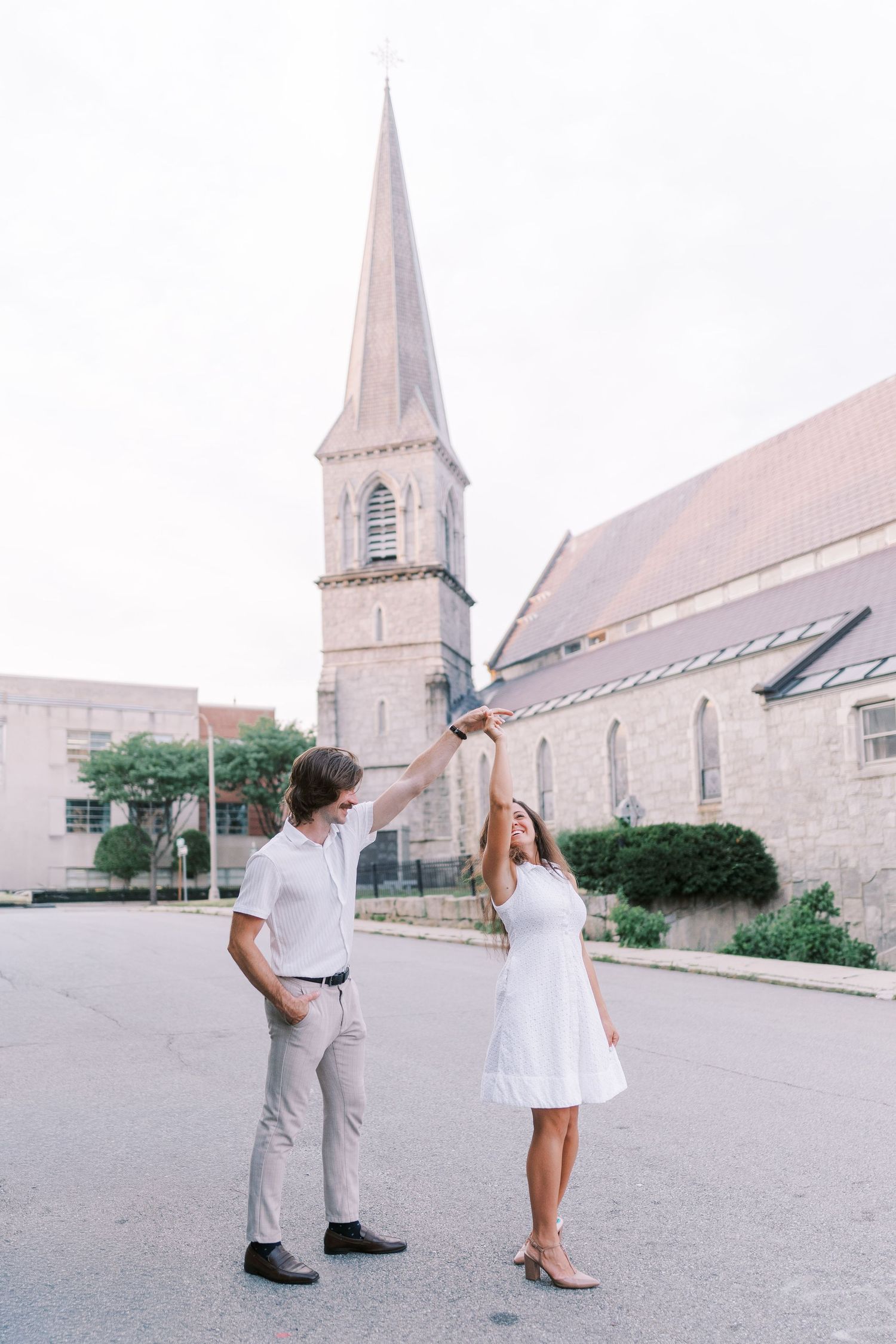 A couple poses playfully outside a stone church with a tall spire.