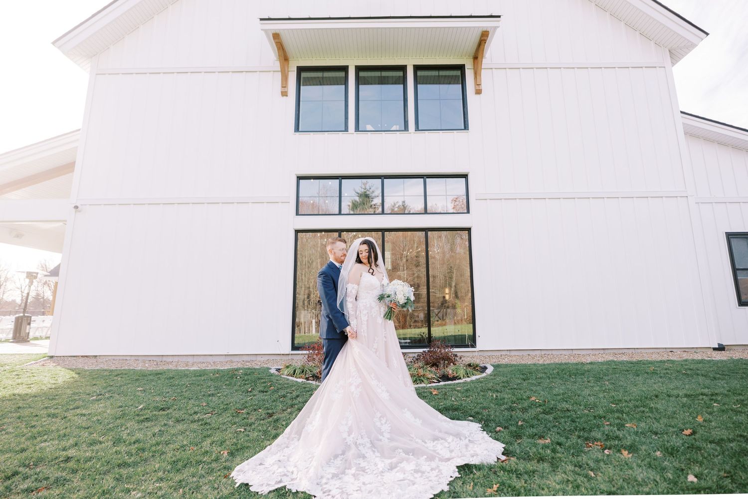 A bride in a flowing white wedding dress poses in front of a white church building with windows.