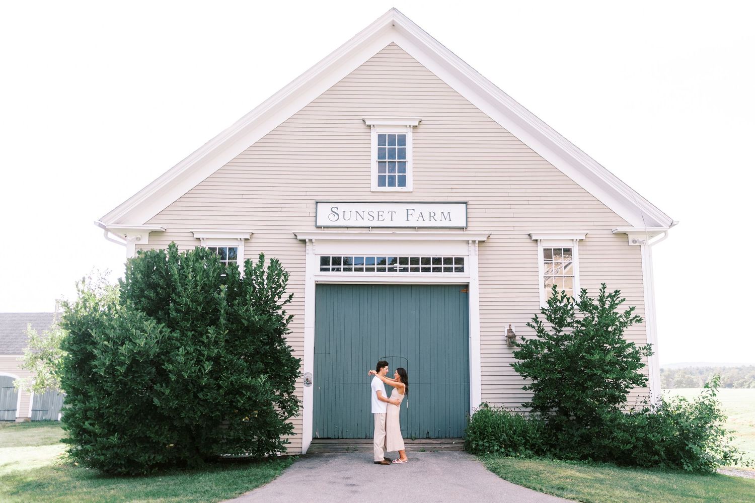 A couple embraces in front of a beige barn with dark green doors and evergreen bushes at the entrance.