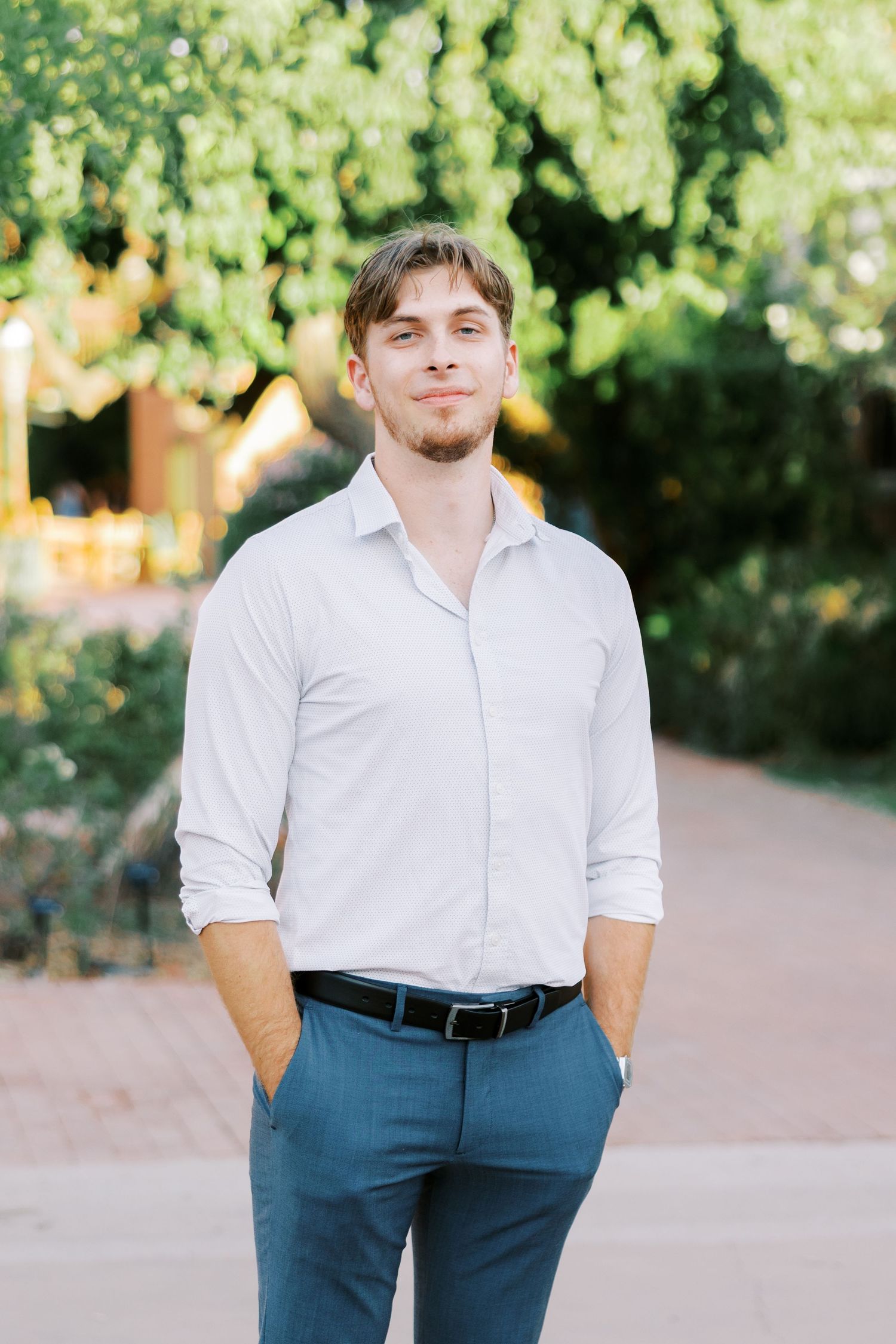 Man in white shirt and navy pants poses casually outdoors on a garden path.