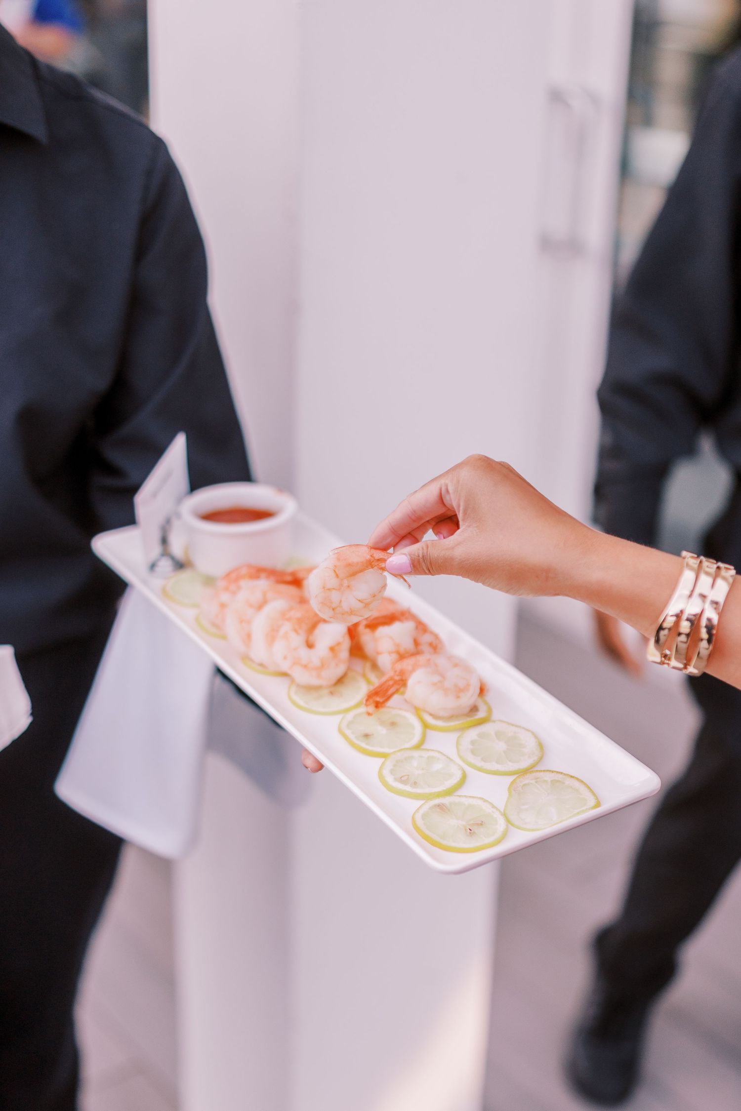 Hands holding white serving trays with shrimp cocktails and lemon slices being passed at an elegant event.
