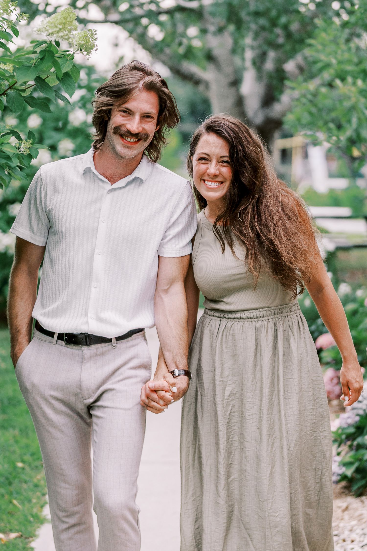 A joyful couple walking outdoors wearing coordinated light-colored outfits holds hands and laughs together in a garden setting.