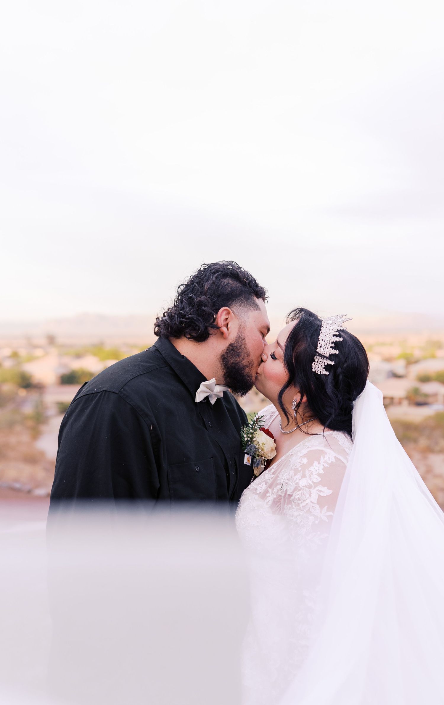 A bride in a white lace gown and veil shares a kiss with their partner against a desert landscape backdrop at sunset.