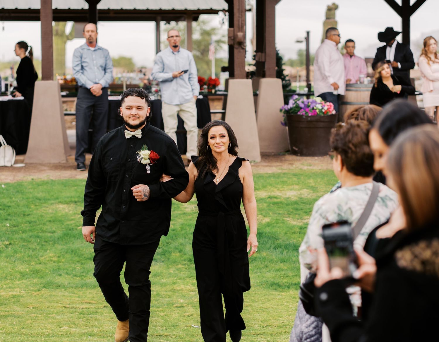A couple dressed in black formal attire walking together at an outdoor wedding reception on a grassy lawn.
